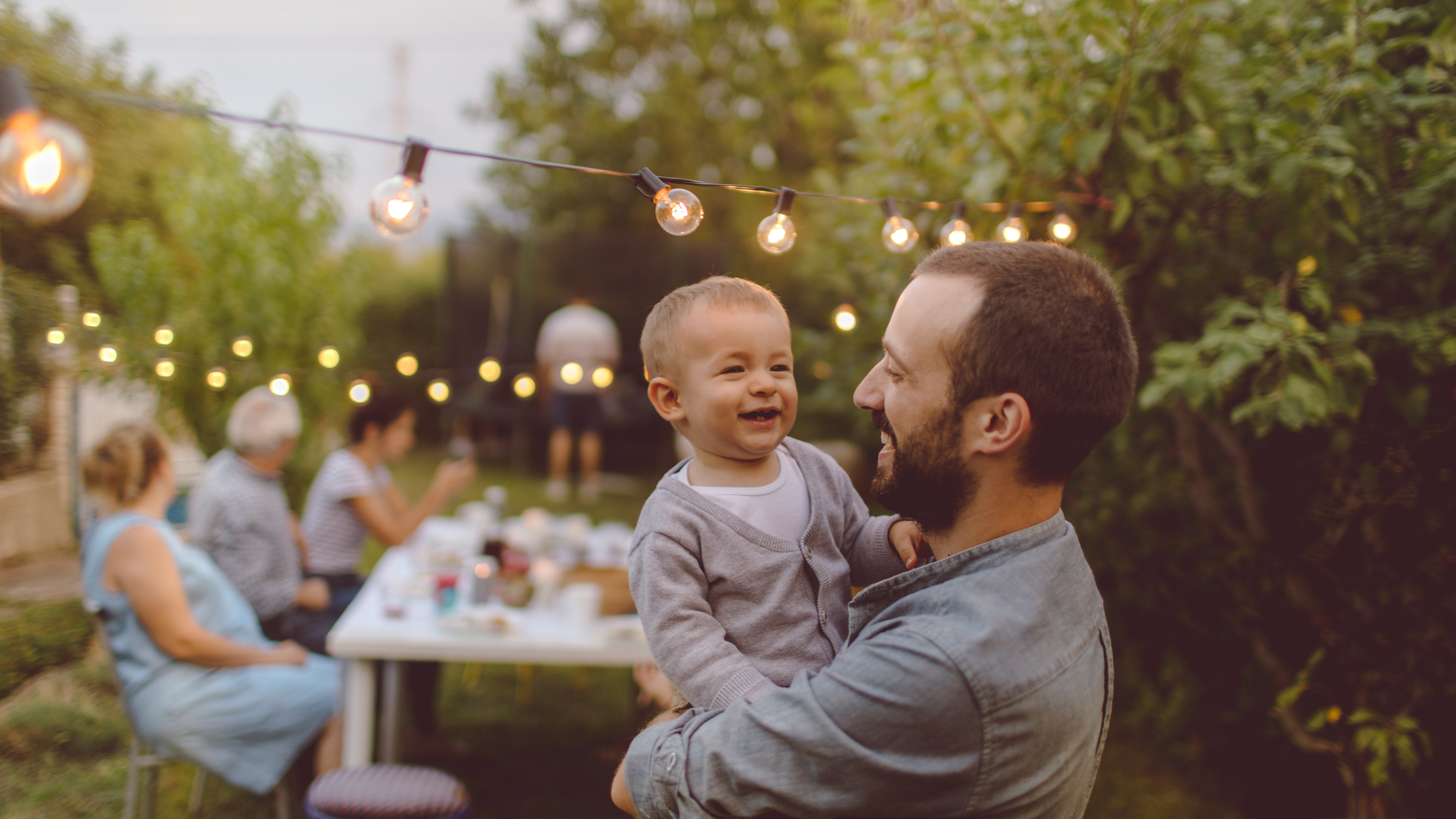 A man is holding a smiling baby in an outdoor setting with string lights and a table in the background.