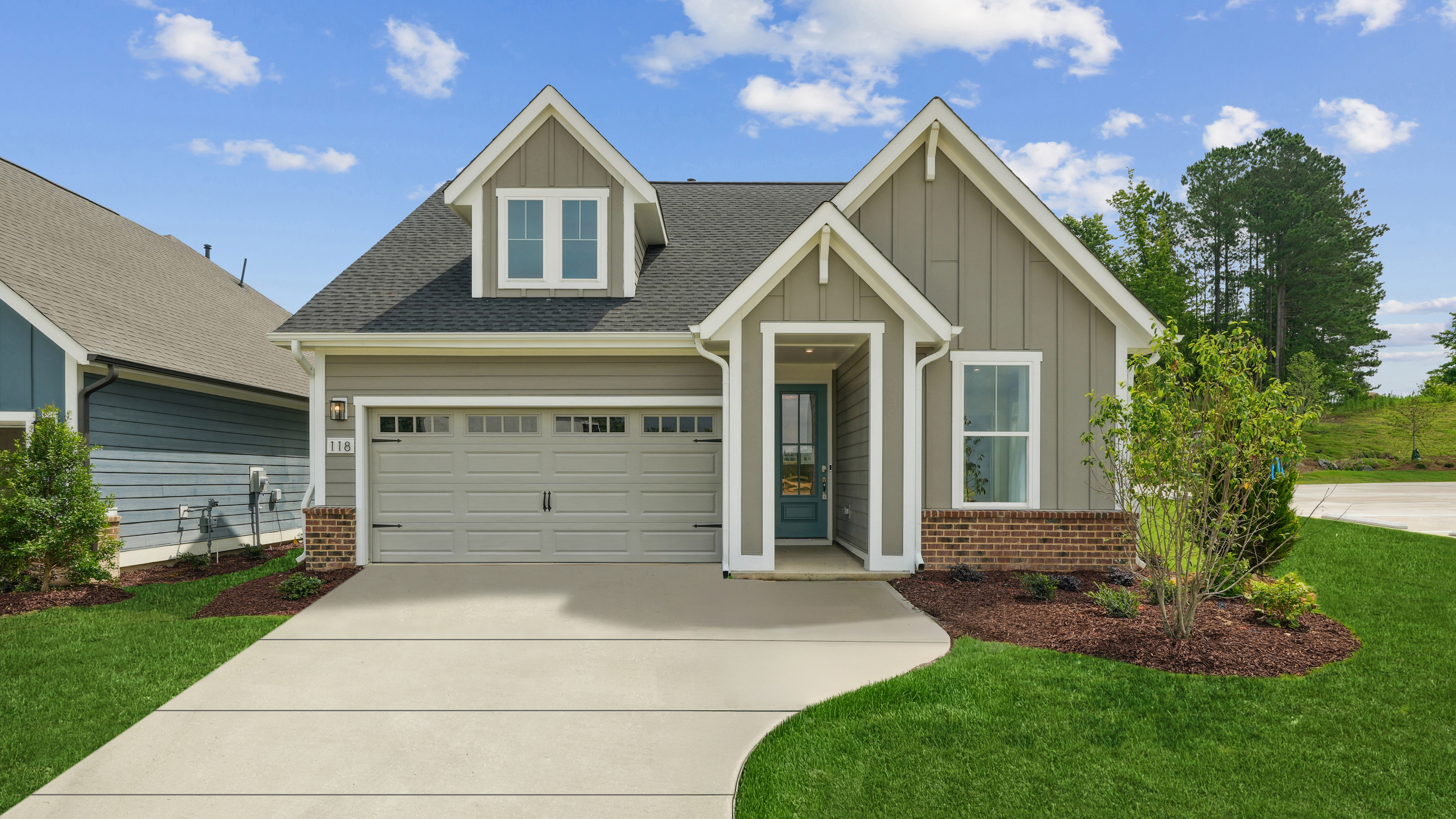 A well-manicured lawn leads to a two-story house with a gray exterior, a garage, and a lush green landscape in the background.