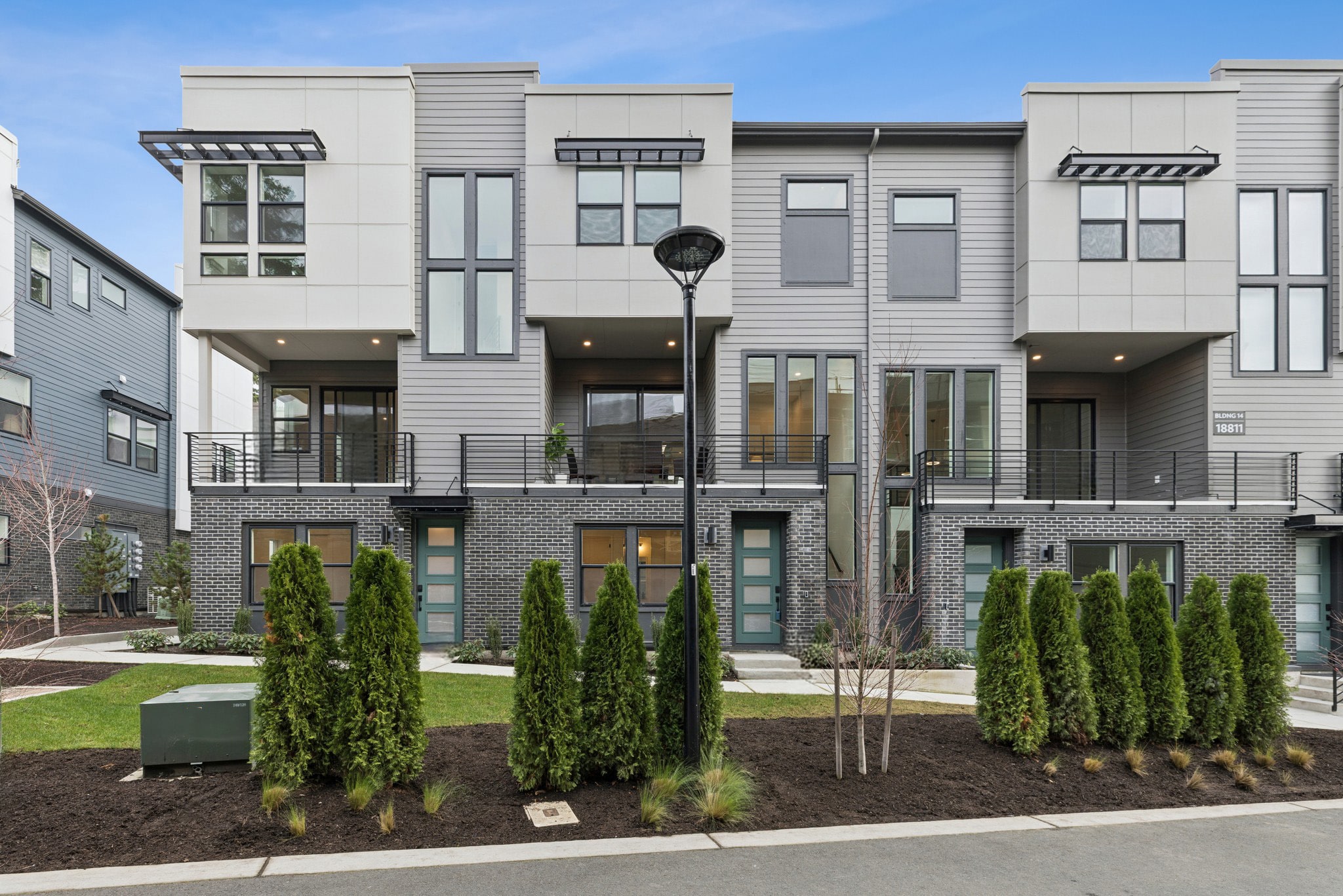 A modern, multi-story residential building with a landscaped front yard featuring evergreen trees and shrubs.