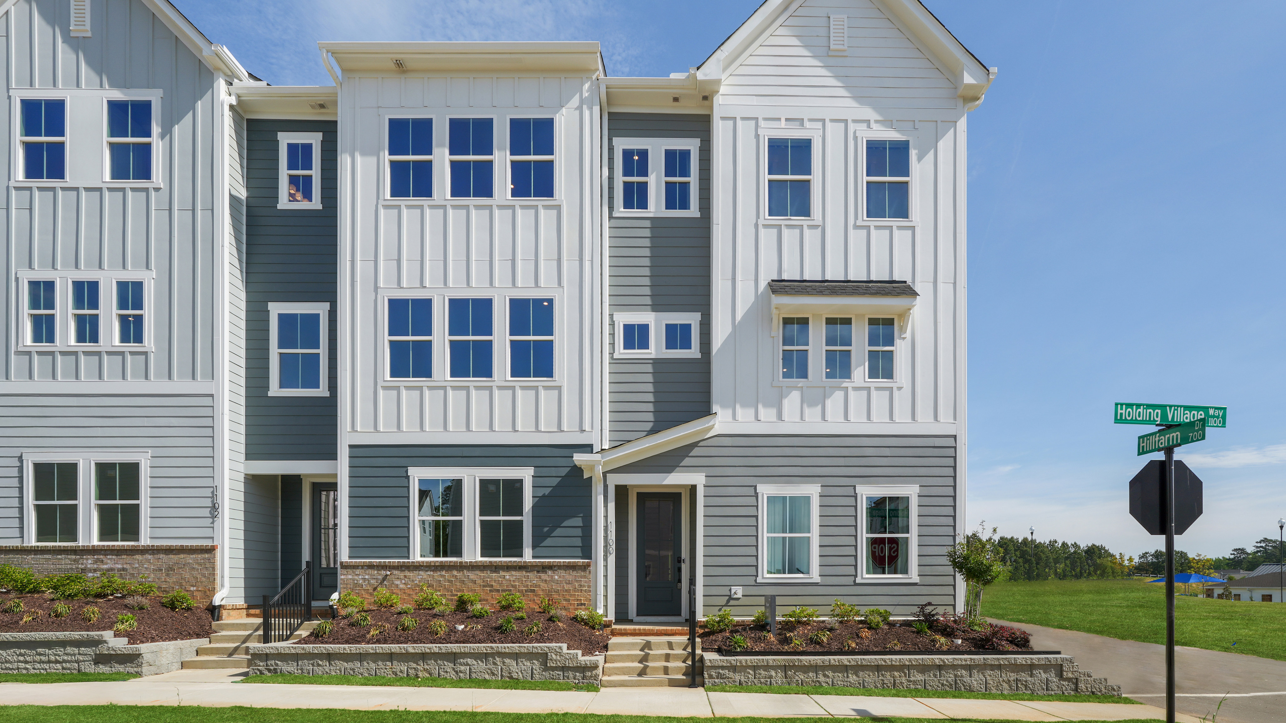 A multi-story residential building with white siding, blue trim, and a landscaped front yard, set against a clear blue sky.