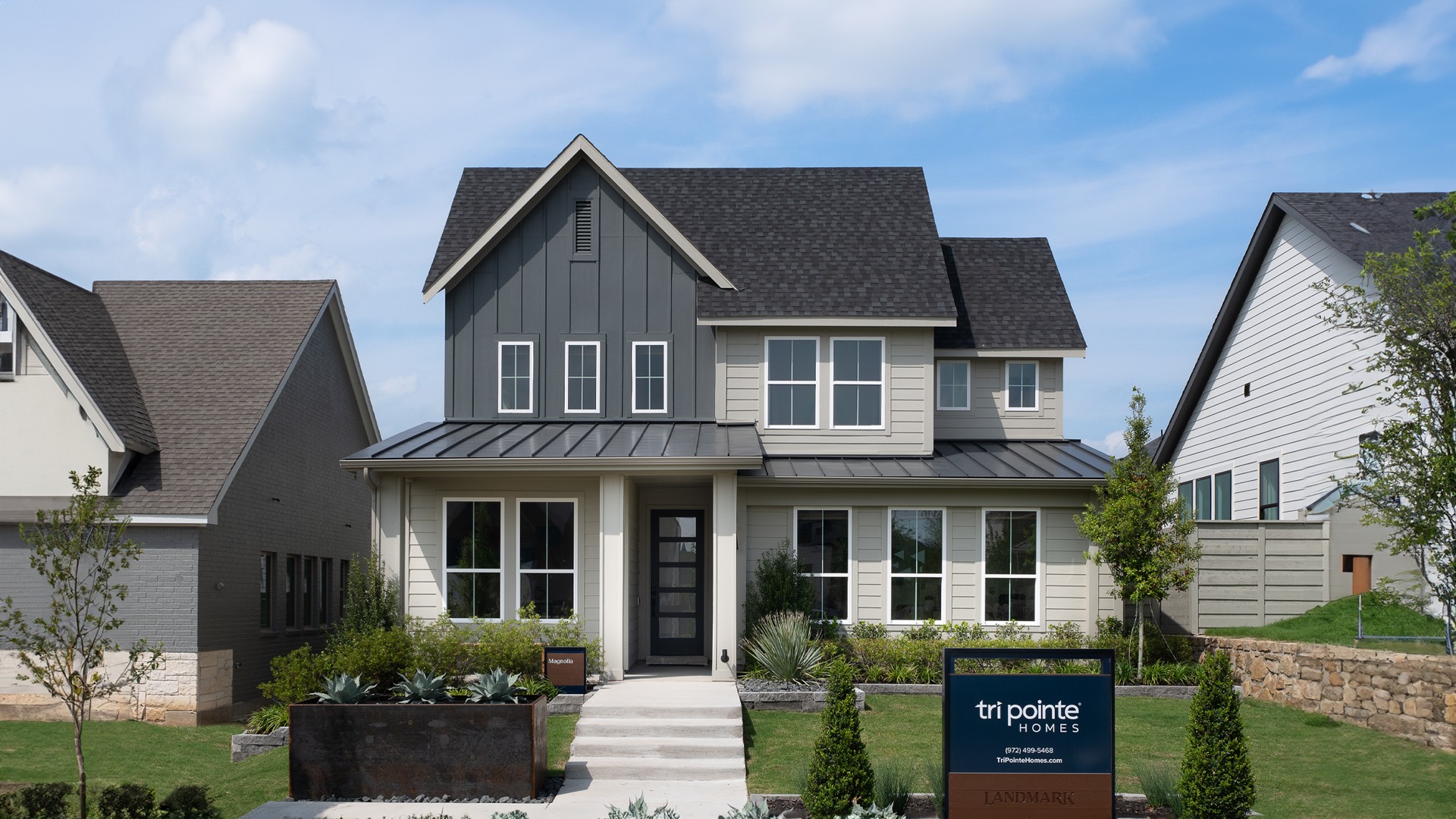 A modern two-story house with a gabled roof, gray siding, and large windows stands in a well-landscaped yard with a small sign in the foreground.