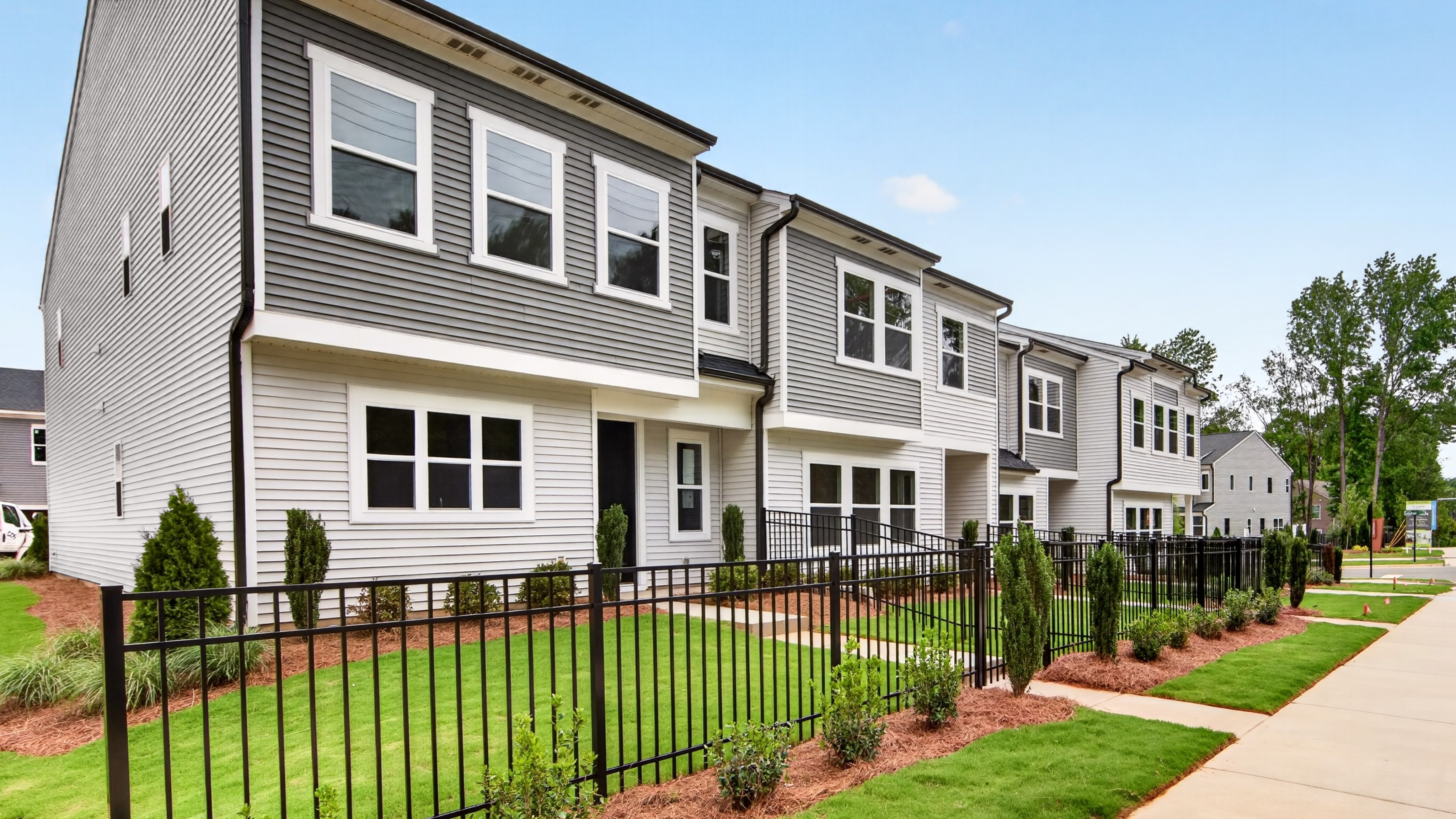 A row of modern townhouses with manicured lawns and black metal fences in the foreground stands against a clear blue sky with scattered trees in the background.