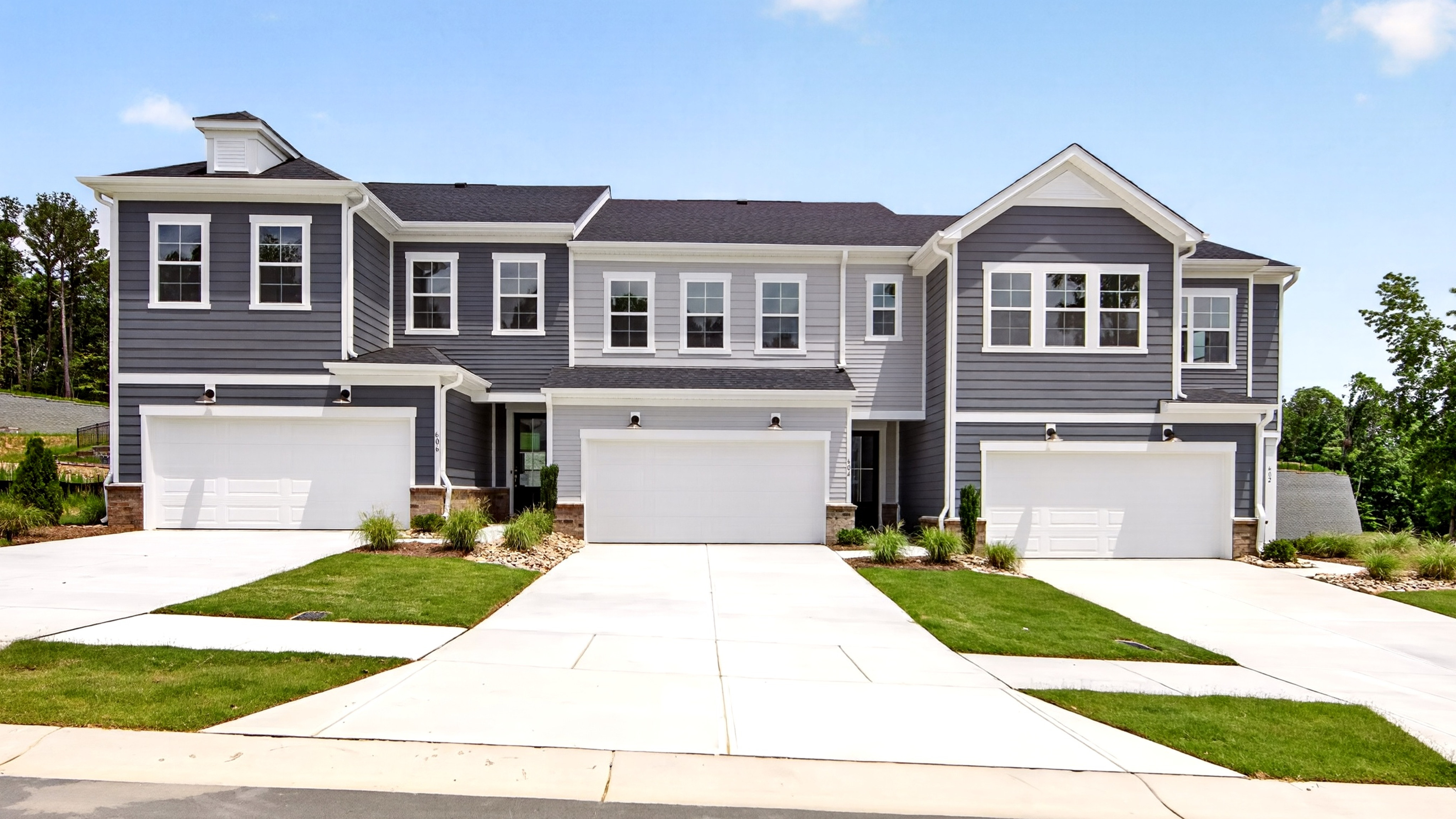 A row of modern townhouses with white garage doors and manicured lawns in the foreground sits against a backdrop of trees and a clear blue sky.