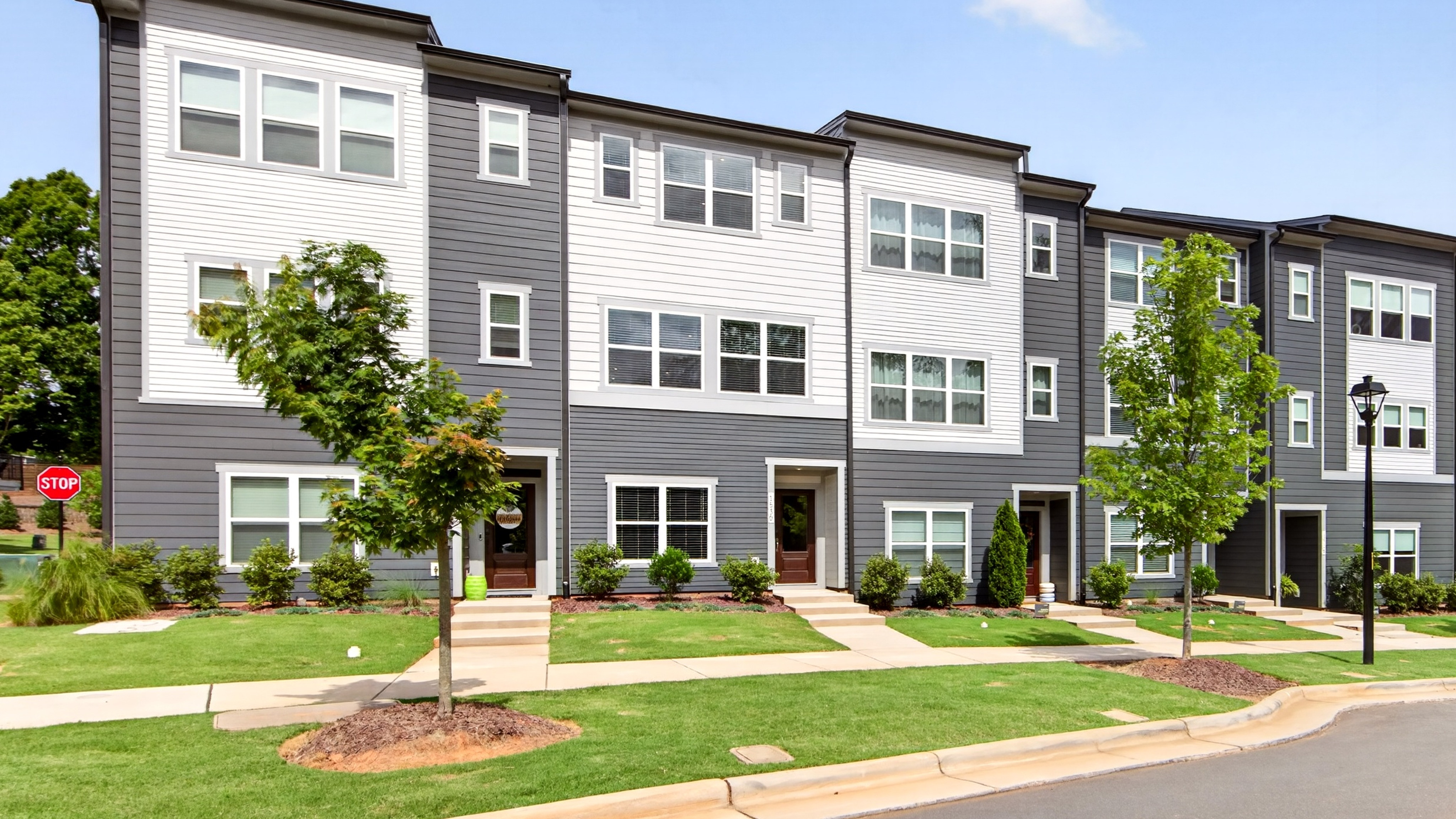 A row of modern townhouses with gray and white exteriors stands on a green lawn with trees in the foreground, against a clear blue sky in the background.