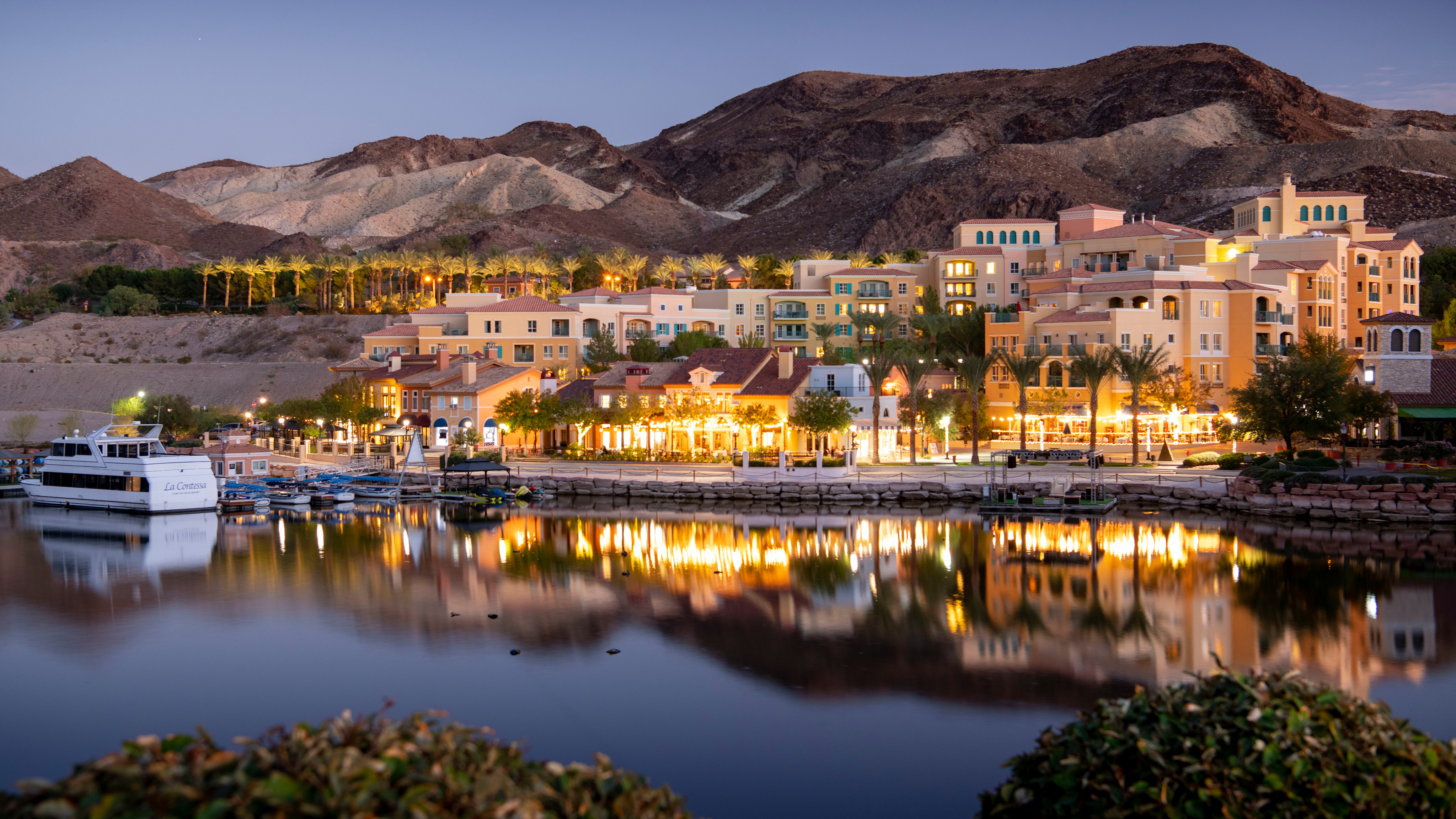 A white boat is docked in a harbor with illuminated buildings reflecting in the calm water, while distant mountains are silhouetted against the twilight sky.