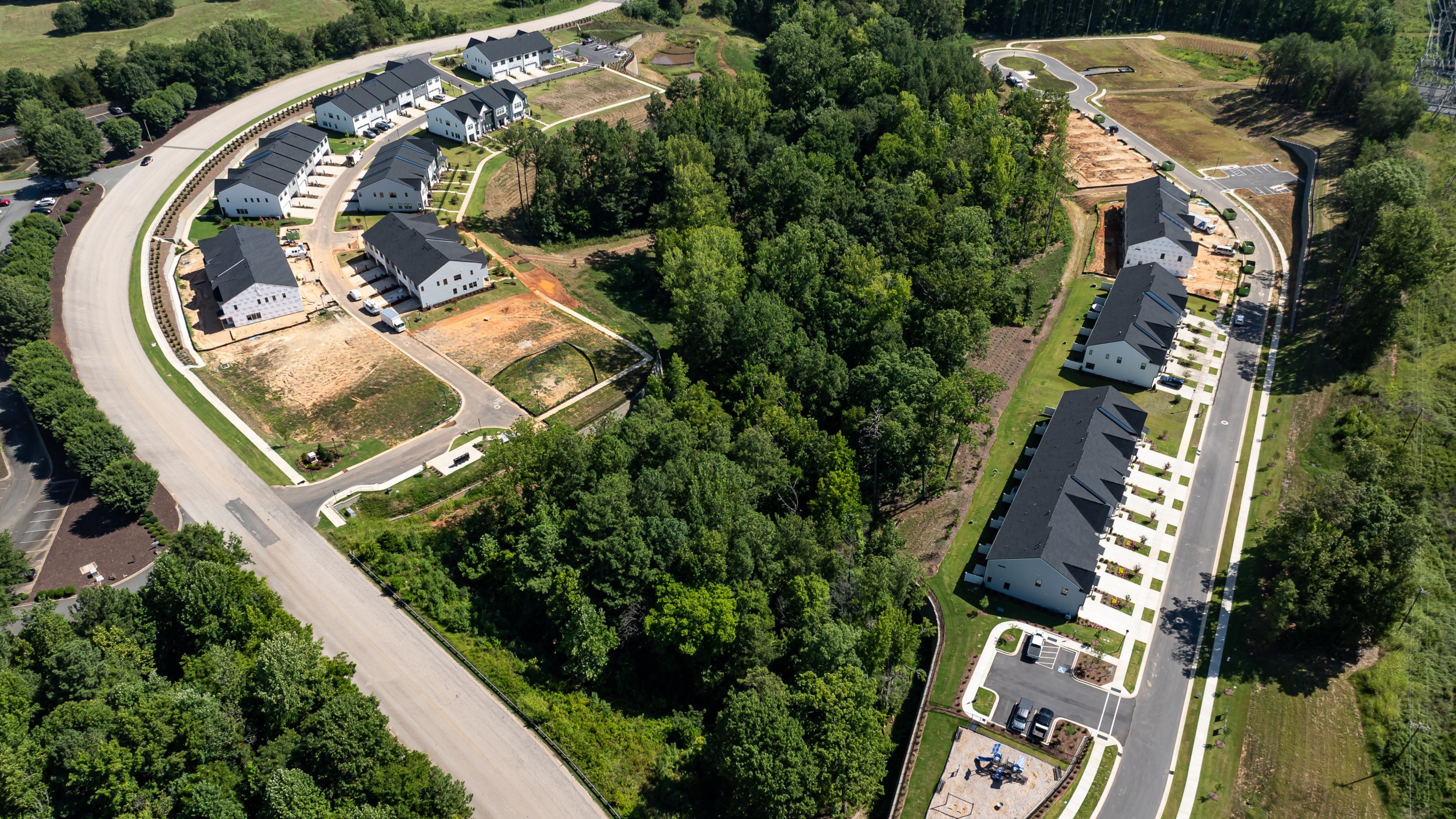 A curved road in the foreground leads to a new housing development with dark-roofed homes nestled among trees and undeveloped land in the background.