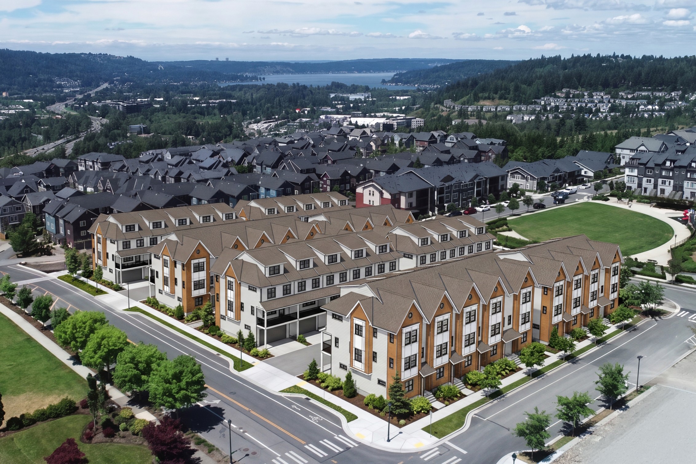 Rows of modern townhouses with light-colored facades and brown roofs fill the foreground, while a sprawling suburban landscape with a distant body of water and tree-covered hills forms the background.