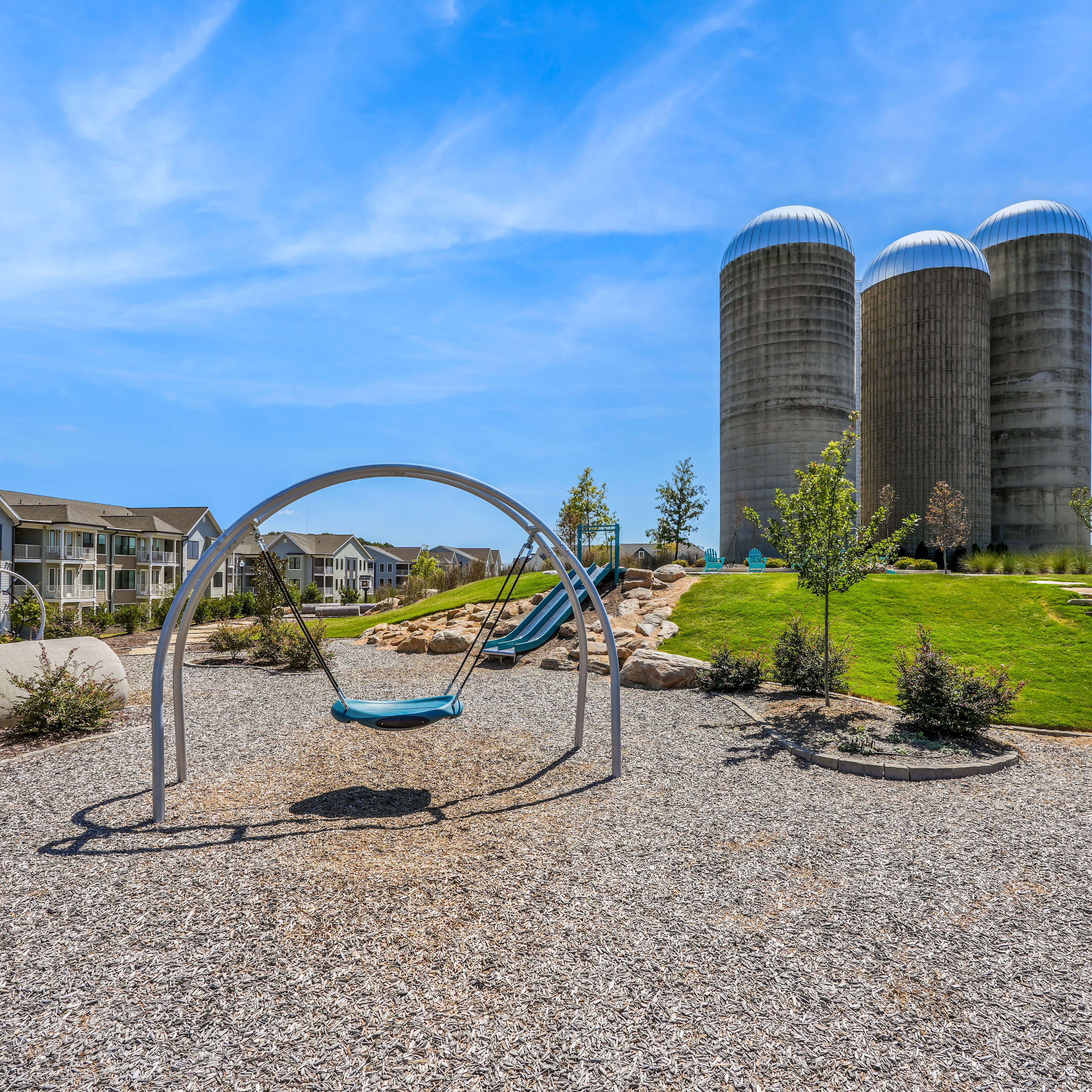 A playground with a swing set sits in the foreground, surrounded by a grassy area and landscaping. In the background, tall silos and residential buildings can be seen against a blue sky with wispy clouds.