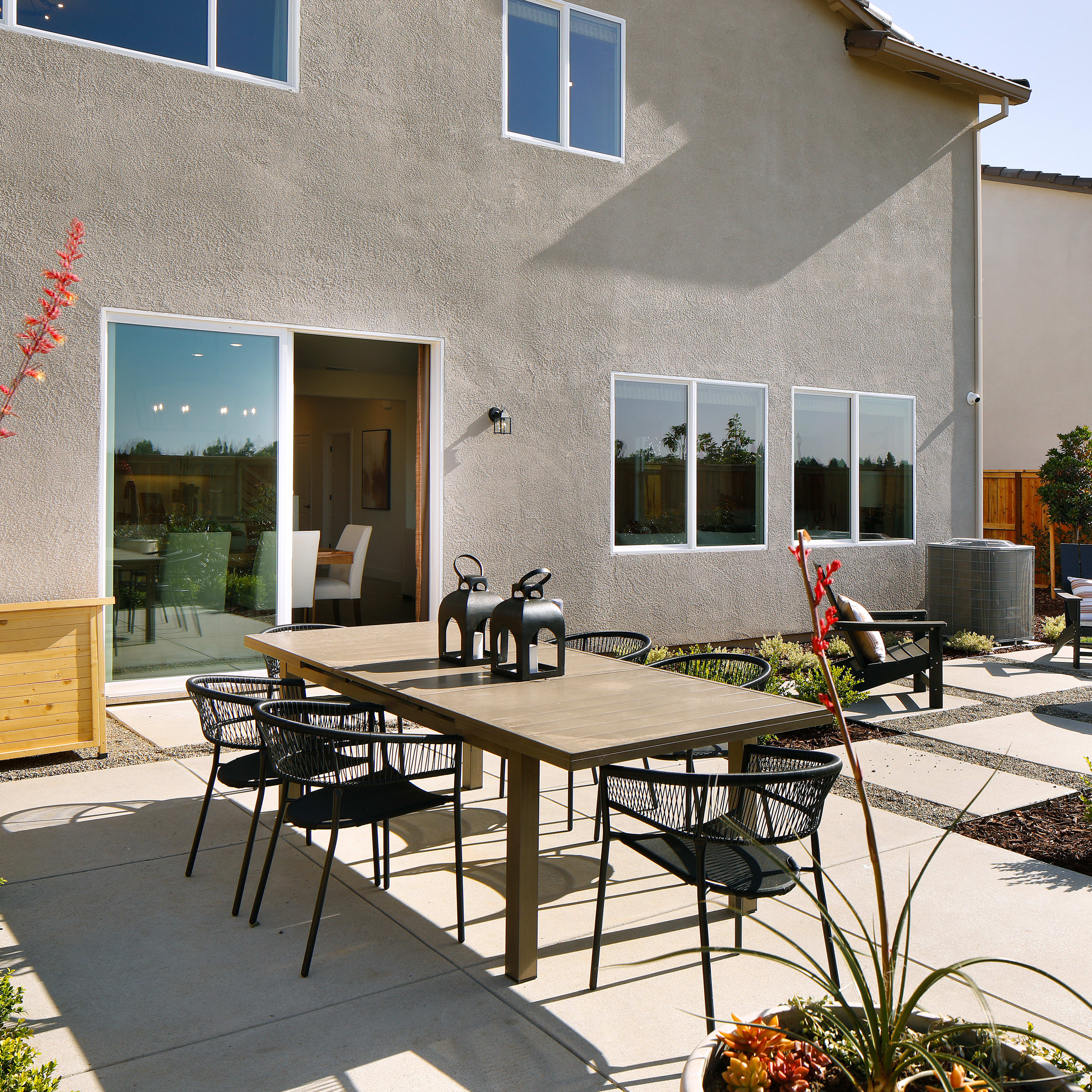 A well-designed outdoor patio area with a dining table and chairs, surrounded by lush greenery and a modern, stucco-clad home in the background.