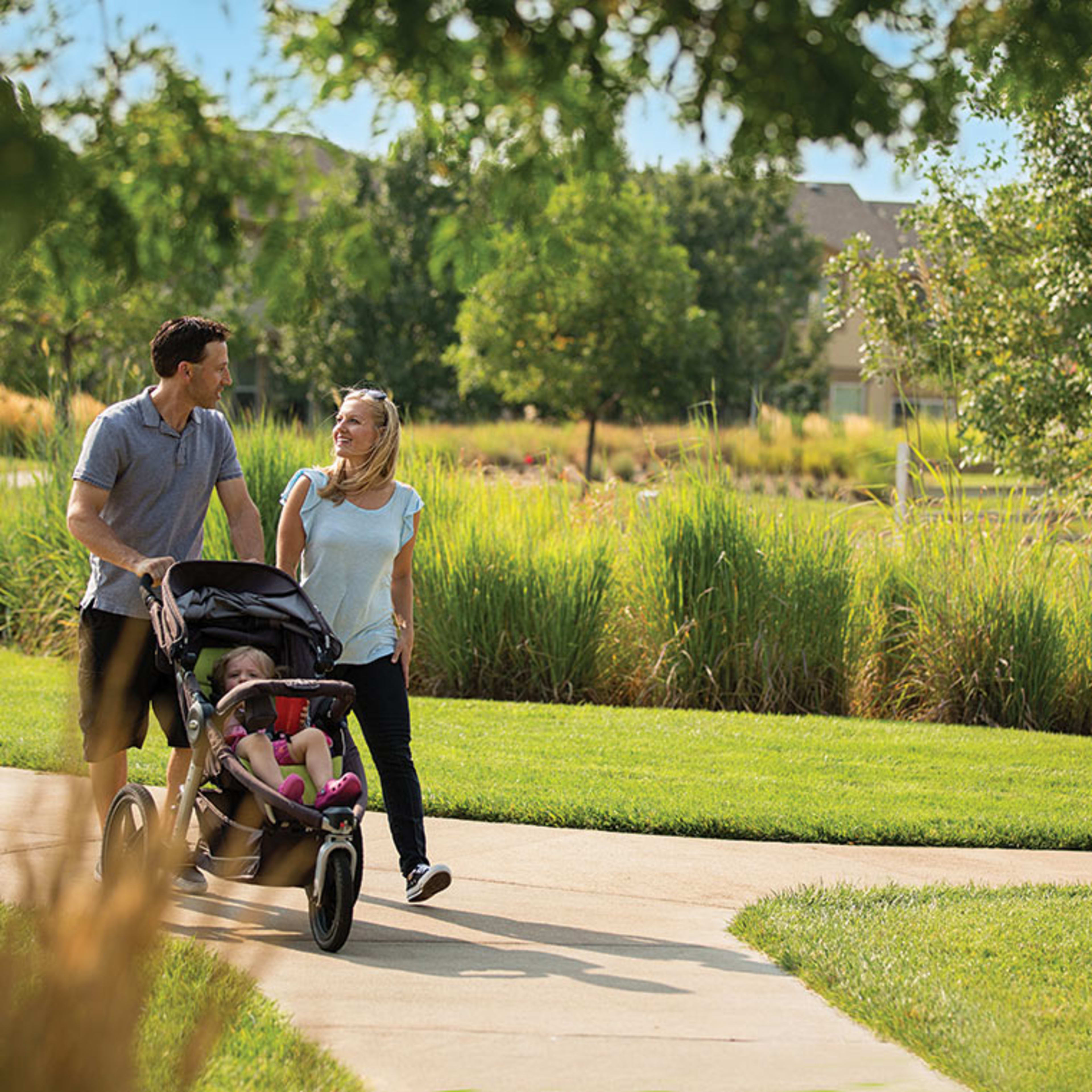 A couple walks together on a paved path, pushing a stroller, surrounded by lush greenery and a well-manicured lawn in the background.
