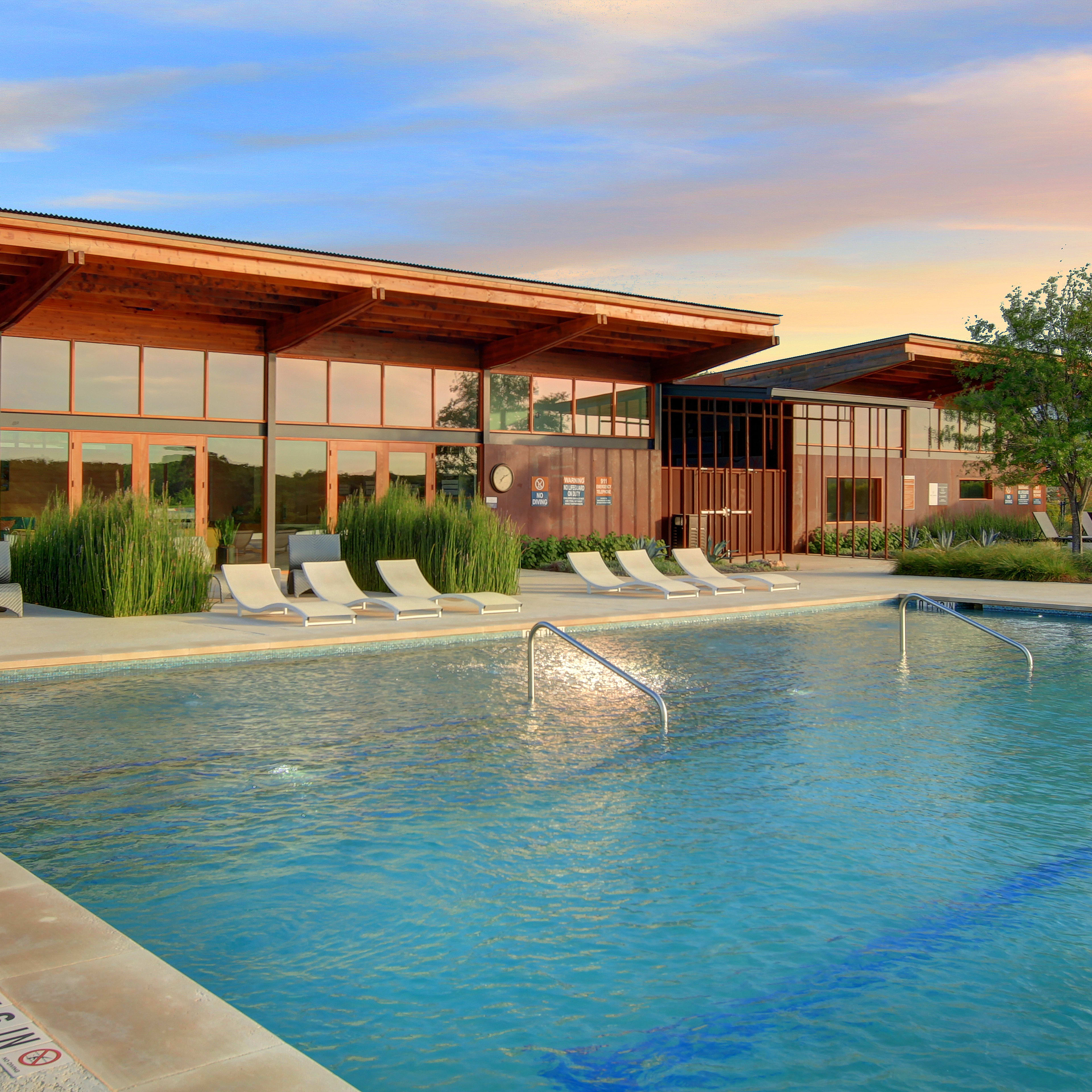 A modern, wooden-framed building with large windows overlooks a serene swimming pool surrounded by lush greenery, set against a vibrant sunset sky.