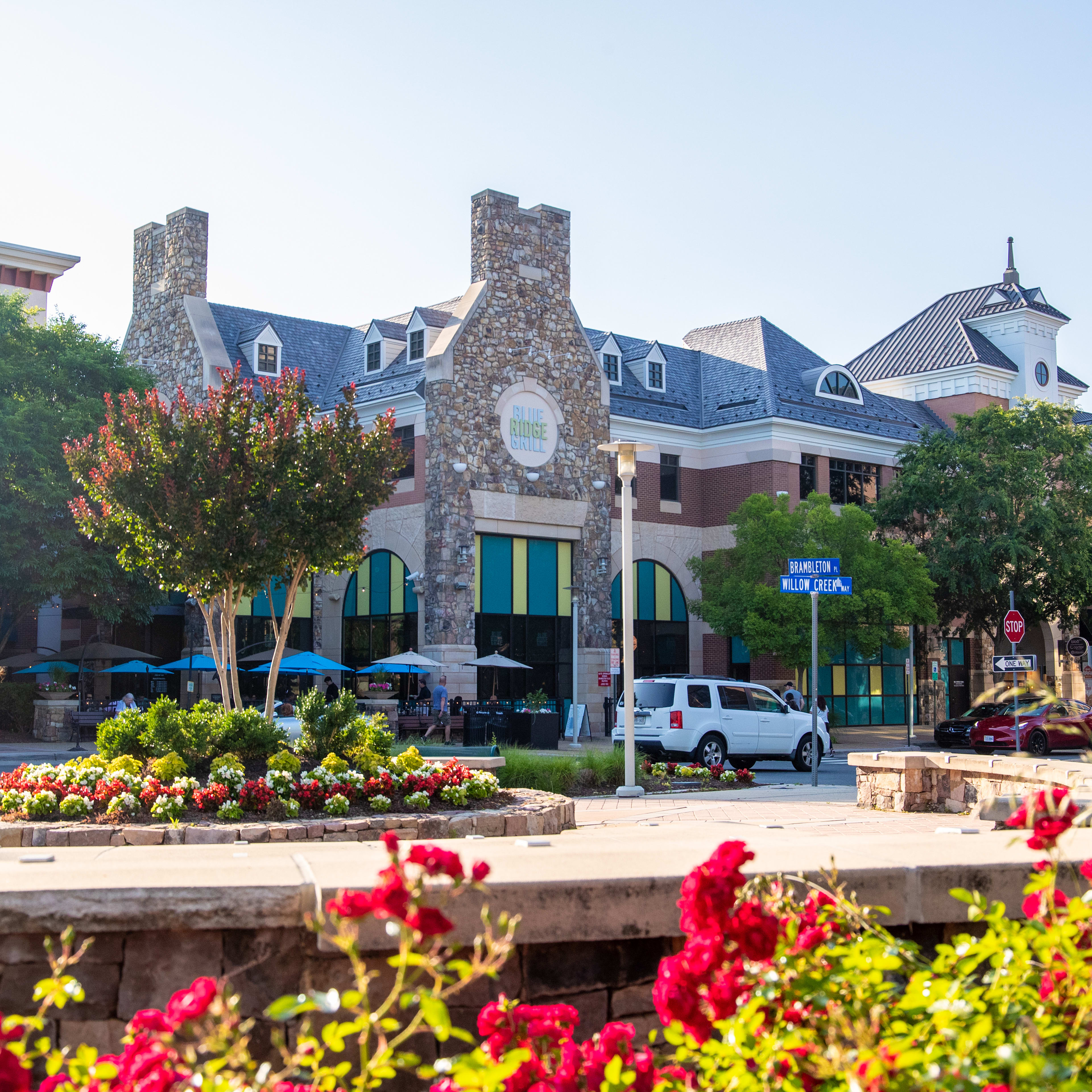 A vibrant and colorful urban scene with a stone fountain surrounded by blooming flowers in the foreground, and a collection of distinctive stone and brick buildings in the background.