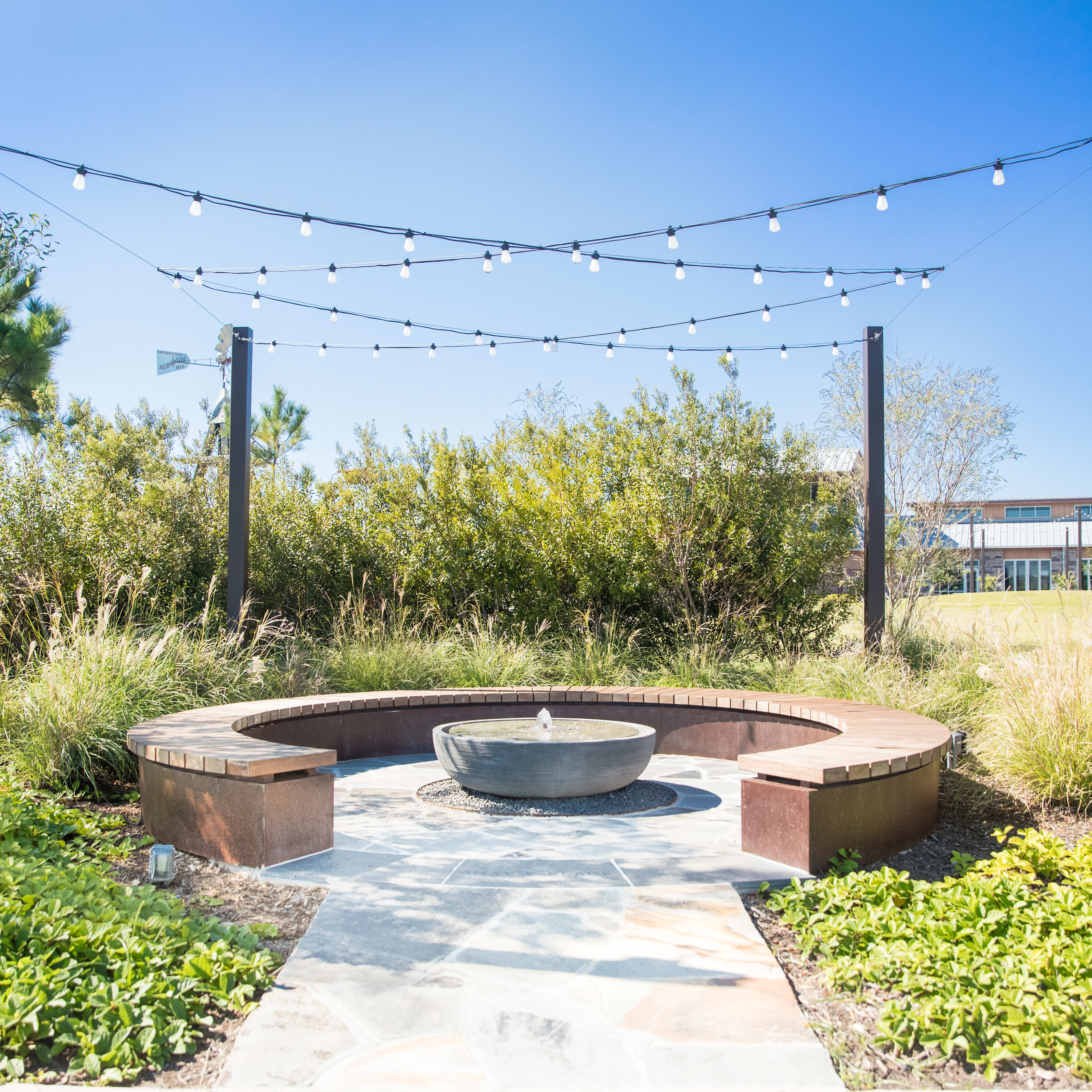 A cozy outdoor seating area with a fire pit surrounded by lush greenery, string lights overhead, and a building visible in the background.