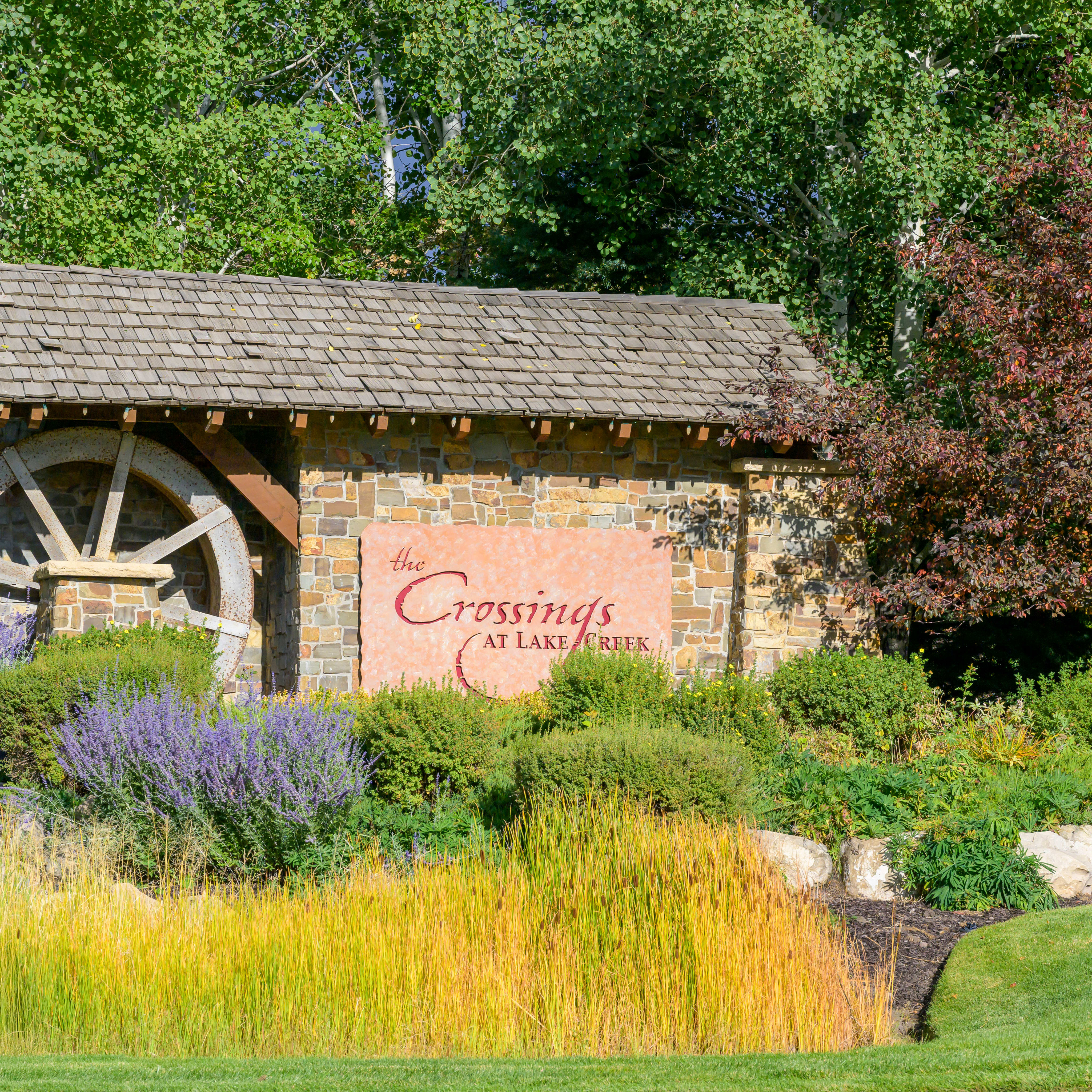 A rustic wooden structure with a water wheel stands amidst a lush, colorful garden, surrounded by a verdant landscape of trees and shrubs.