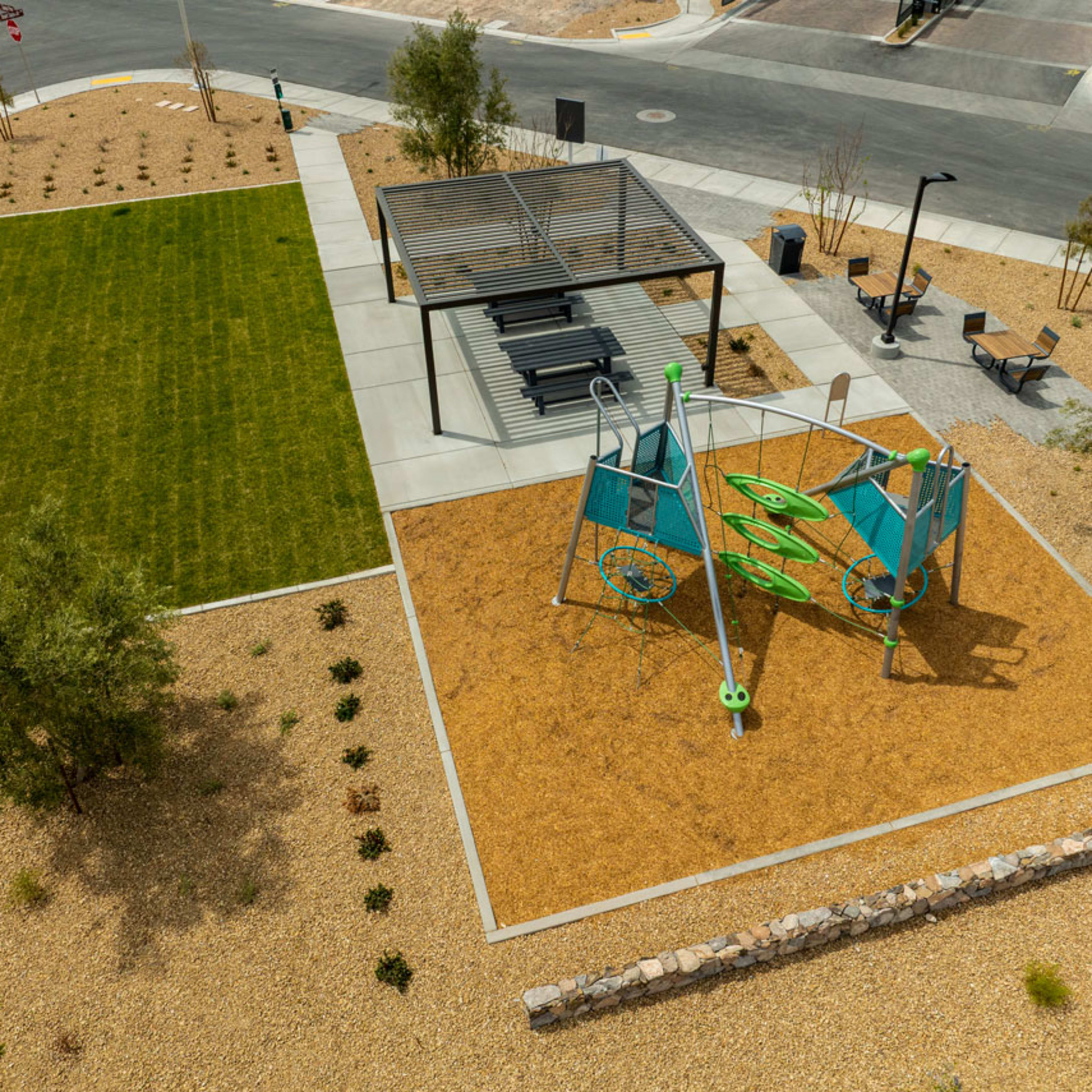 A vibrant outdoor public space with a grassy area, a colorful playground, and various seating options surrounded by paved walkways and landscaping.