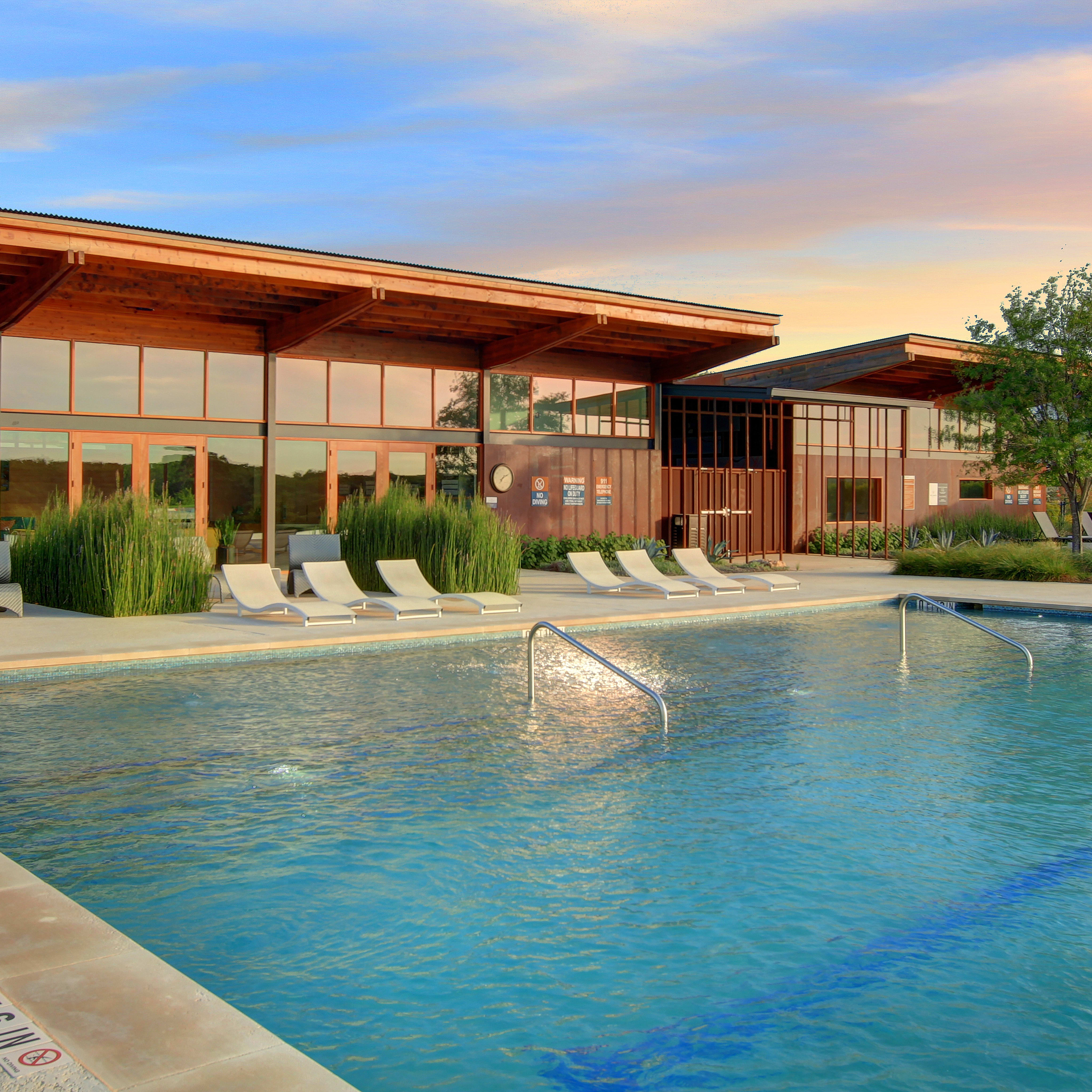 A modern, wooden-framed building with large windows overlooks a serene swimming pool surrounded by lush greenery, set against a vibrant sunset sky.