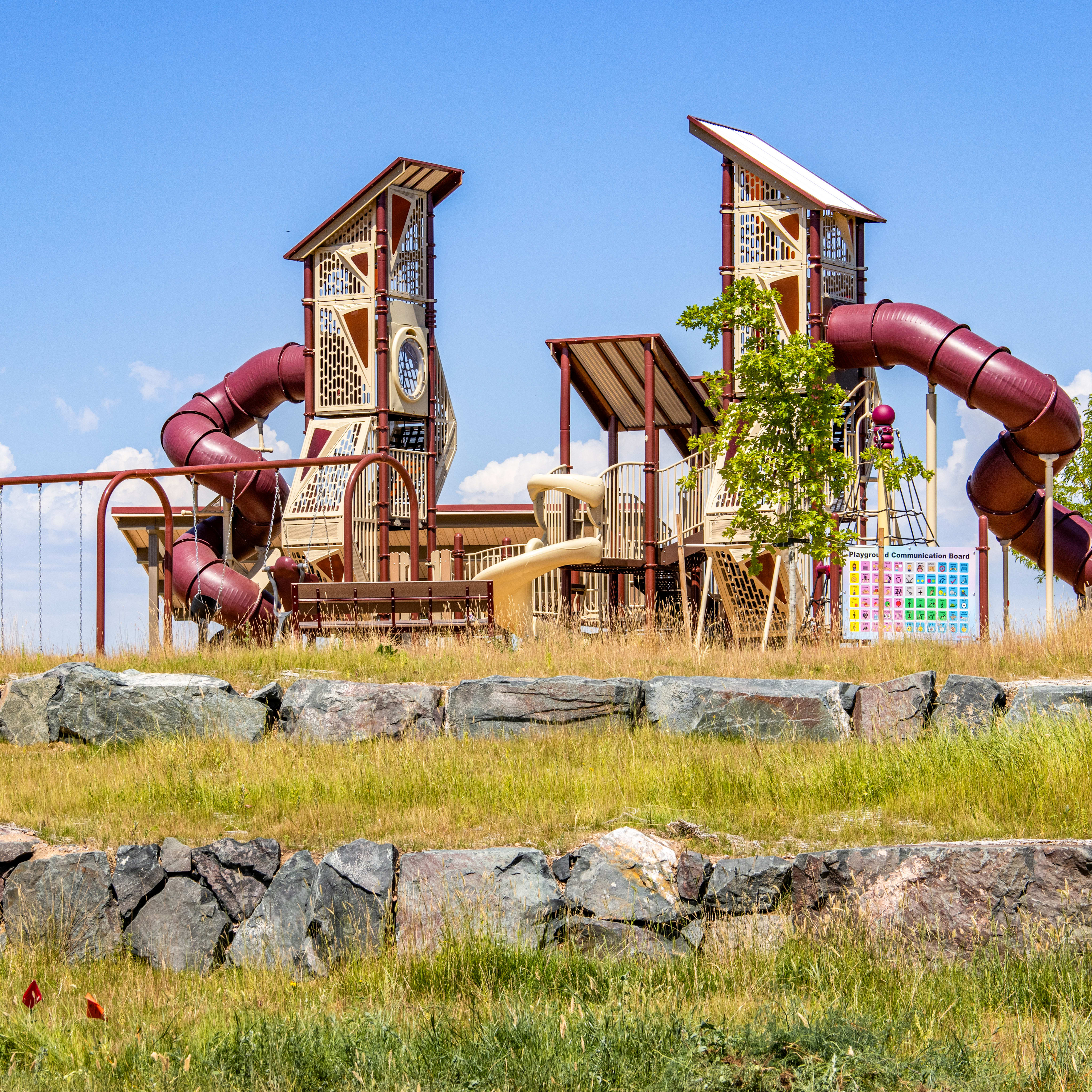 A vibrant playground with slides and swings stands on a grassy hill, with a clear blue sky overhead.
