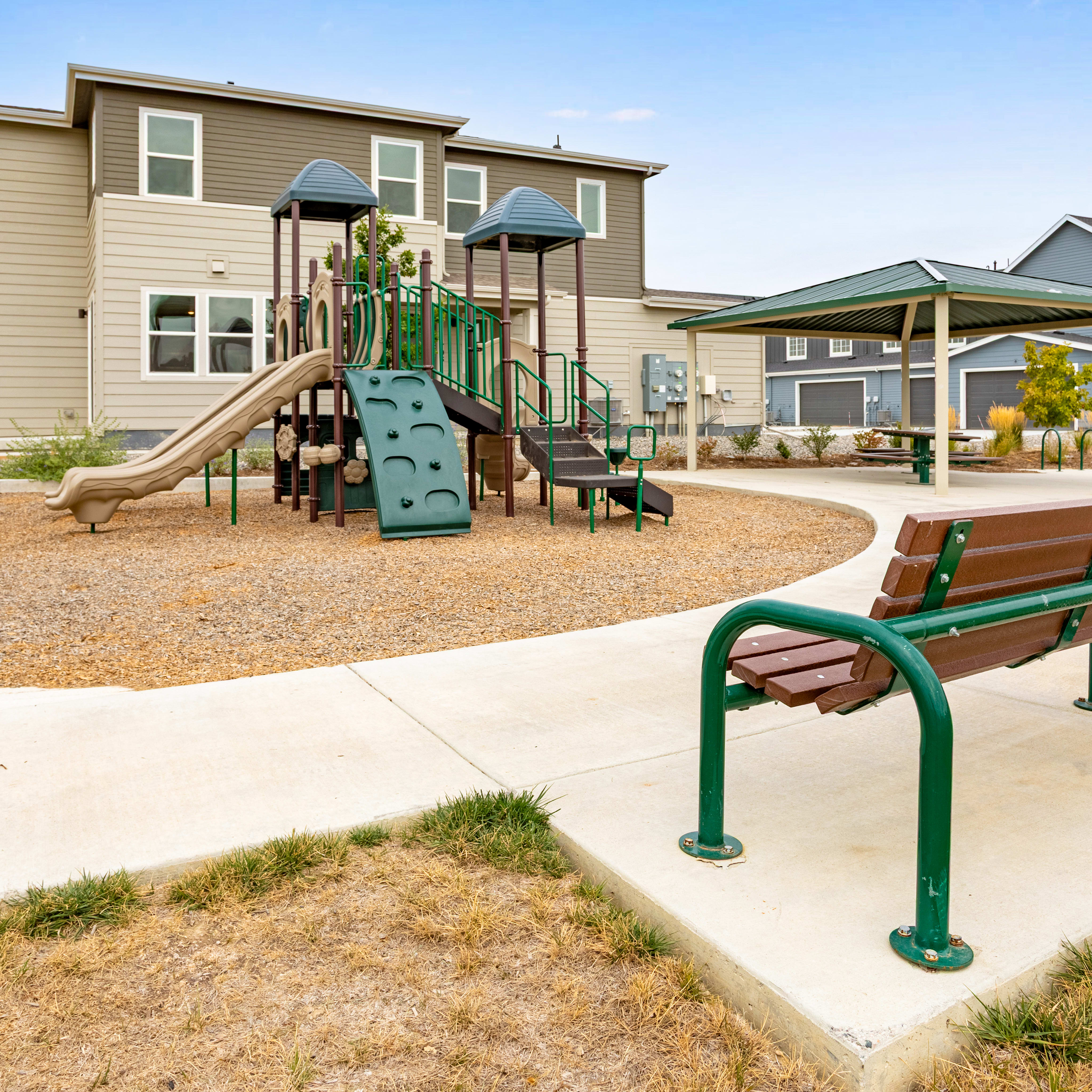 A brown wooden bench with green metal arms sits in the foreground of a playground featuring a climbing structure and a shaded pavilion, with modern residential buildings in the background.