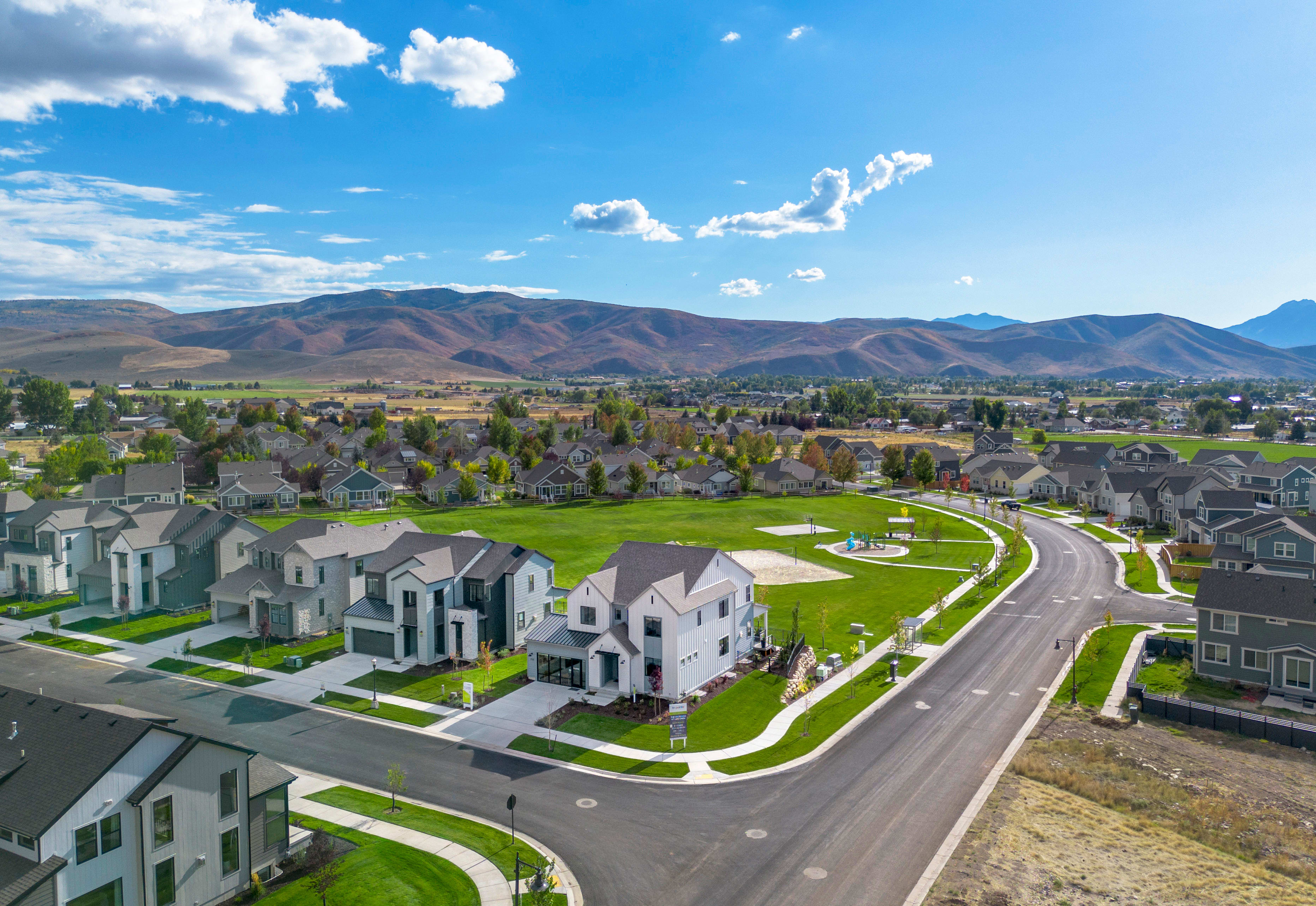 A residential neighborhood nestled in a valley, with rows of houses and a winding road leading through the community, surrounded by rugged mountains and a vibrant blue sky with fluffy white clouds.