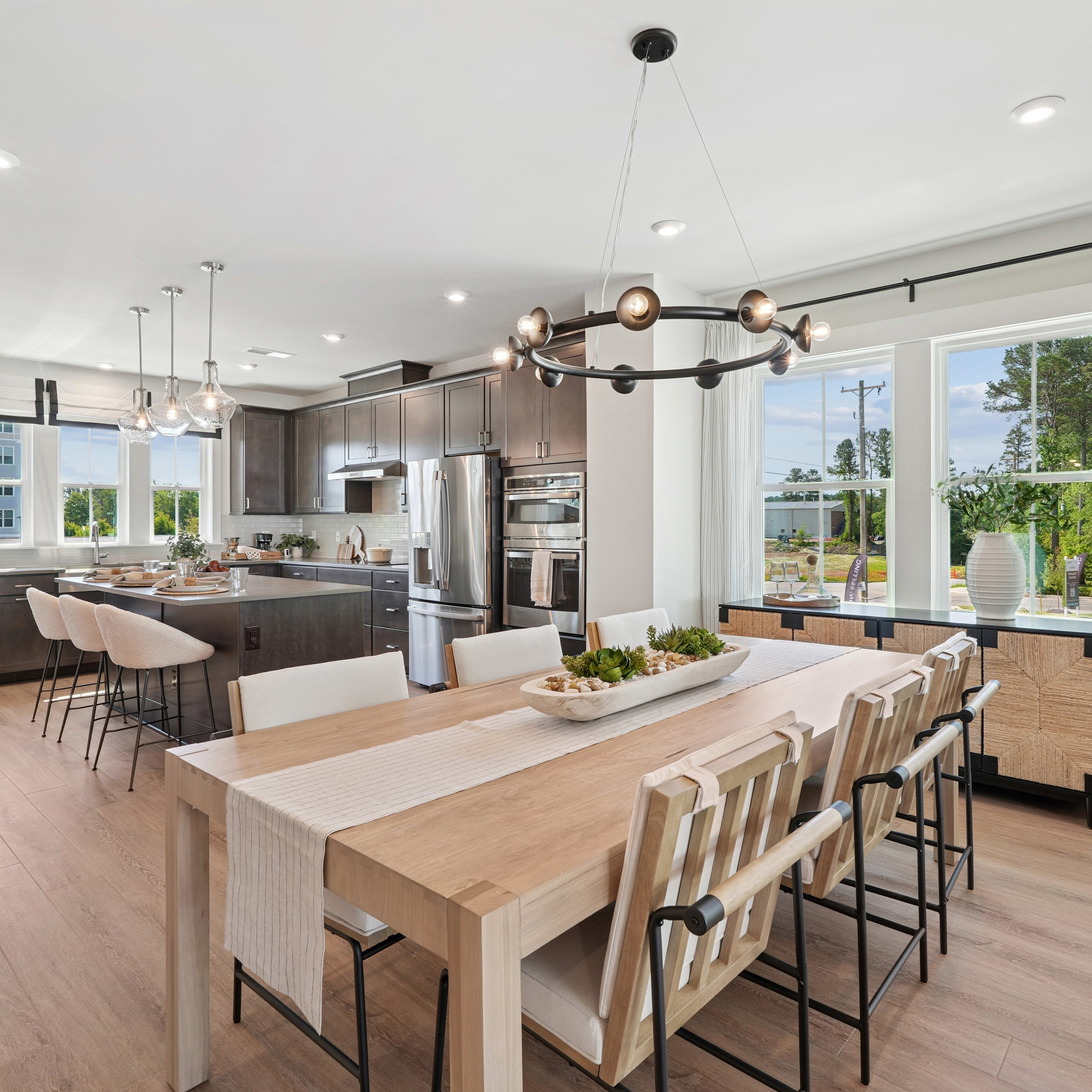 A modern and spacious kitchen with a large wooden dining table, sleek dark cabinets, and a chandelier overhead, surrounded by large windows overlooking a lush outdoor landscape.