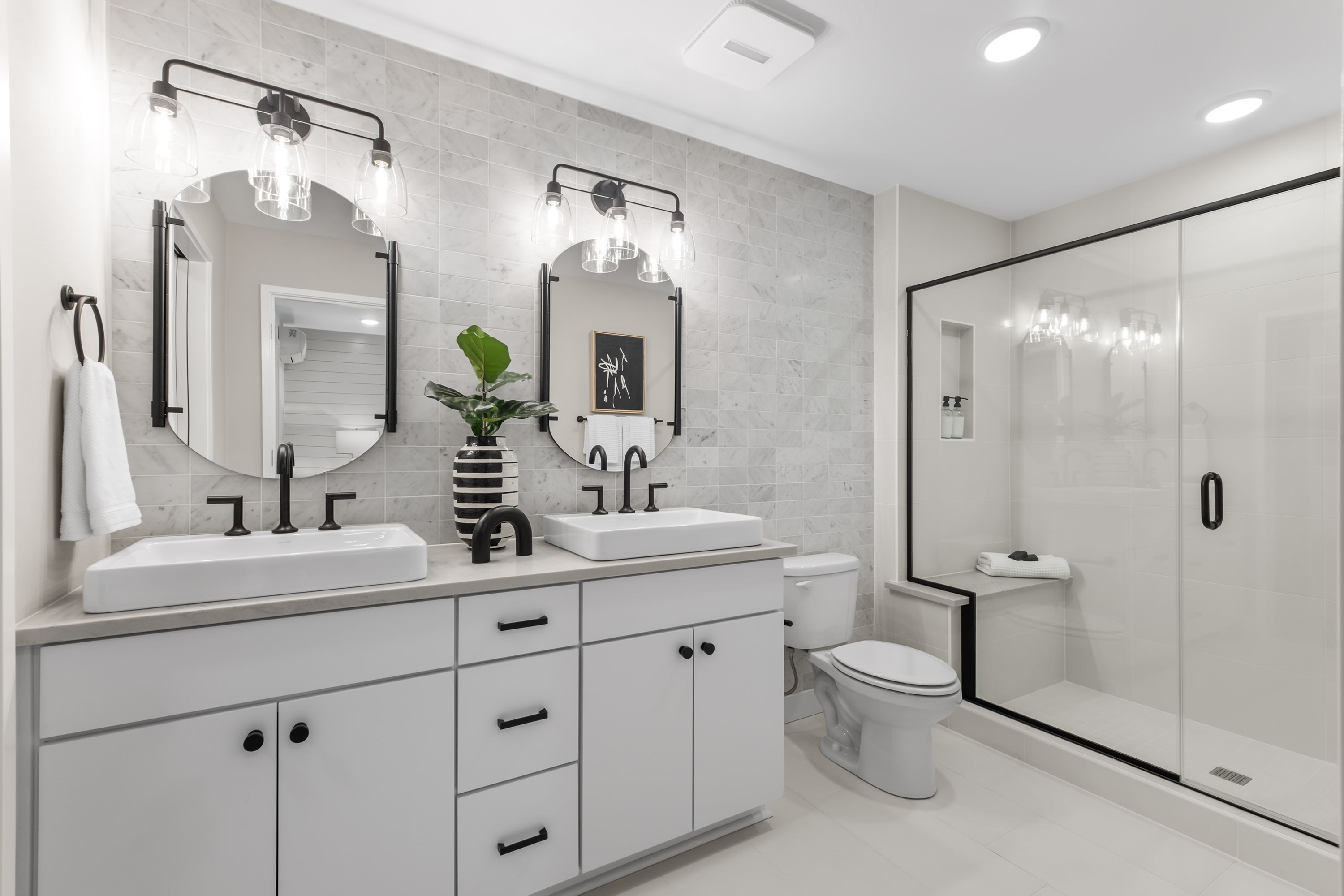 A modern, minimalist bathroom with white cabinets, a double vanity, and a large mirror above it, complemented by black accents and a glass shower enclosure.