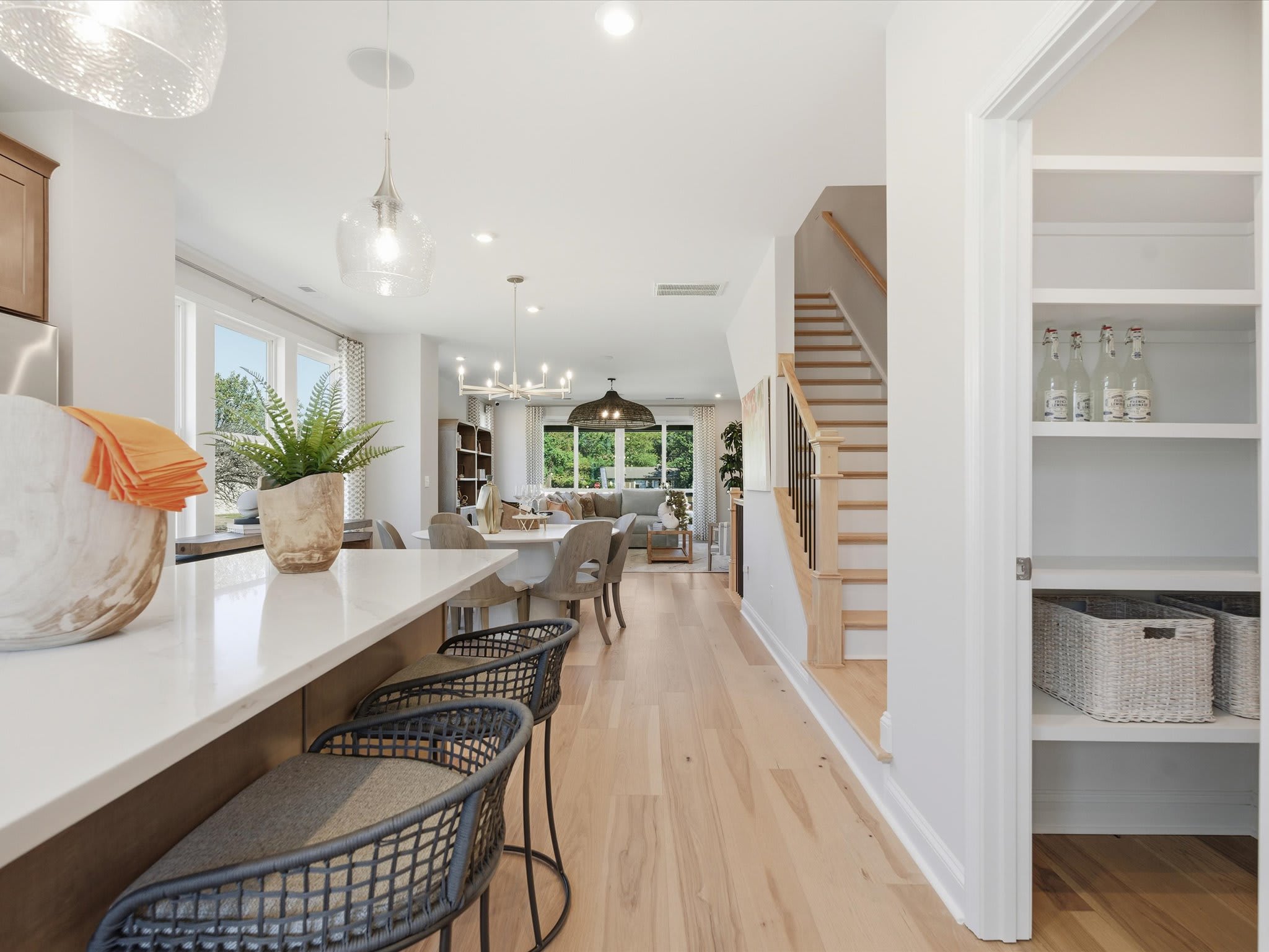 A bright and airy open-concept living space with a long wooden dining table, wicker chairs, and a staircase leading to an upper level.