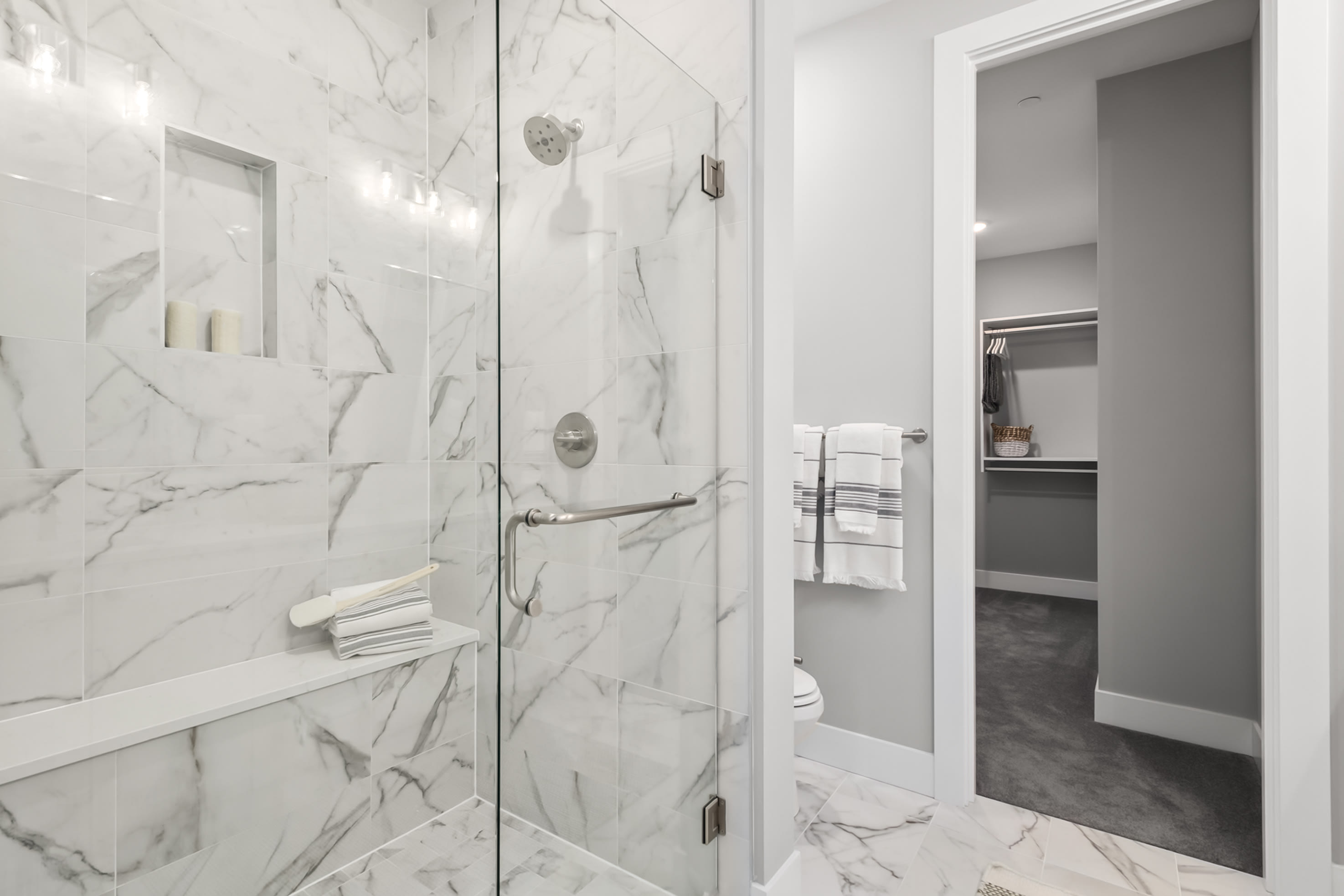 A modern, minimalist bathroom with sleek white marble walls, a glass shower enclosure, and a glimpse of an adjacent room visible through an open doorway.