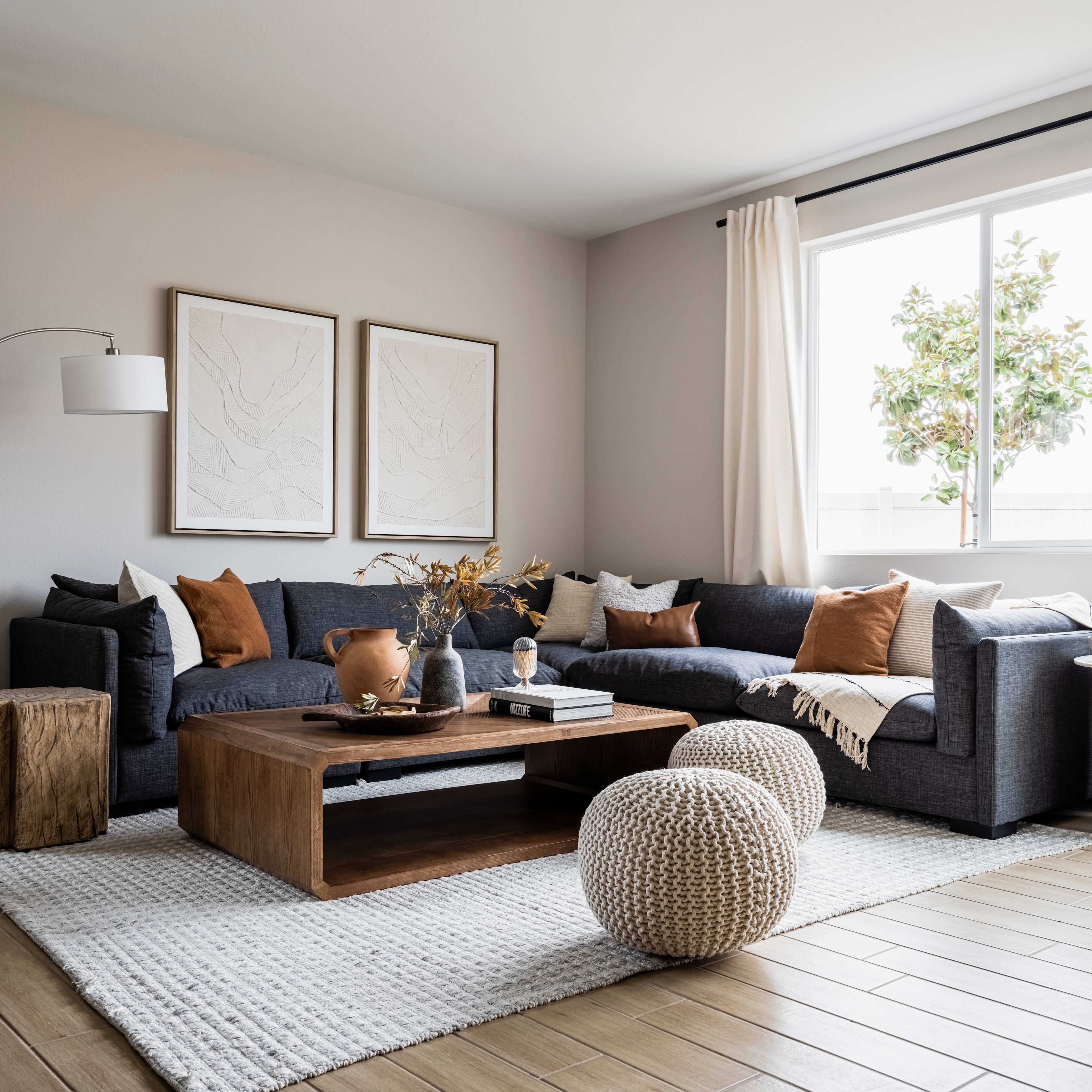 A cozy and inviting living room with a large sectional sofa, wooden coffee table, and decorative accents like throw pillows and a pouf, set against a backdrop of large windows overlooking greenery outside.