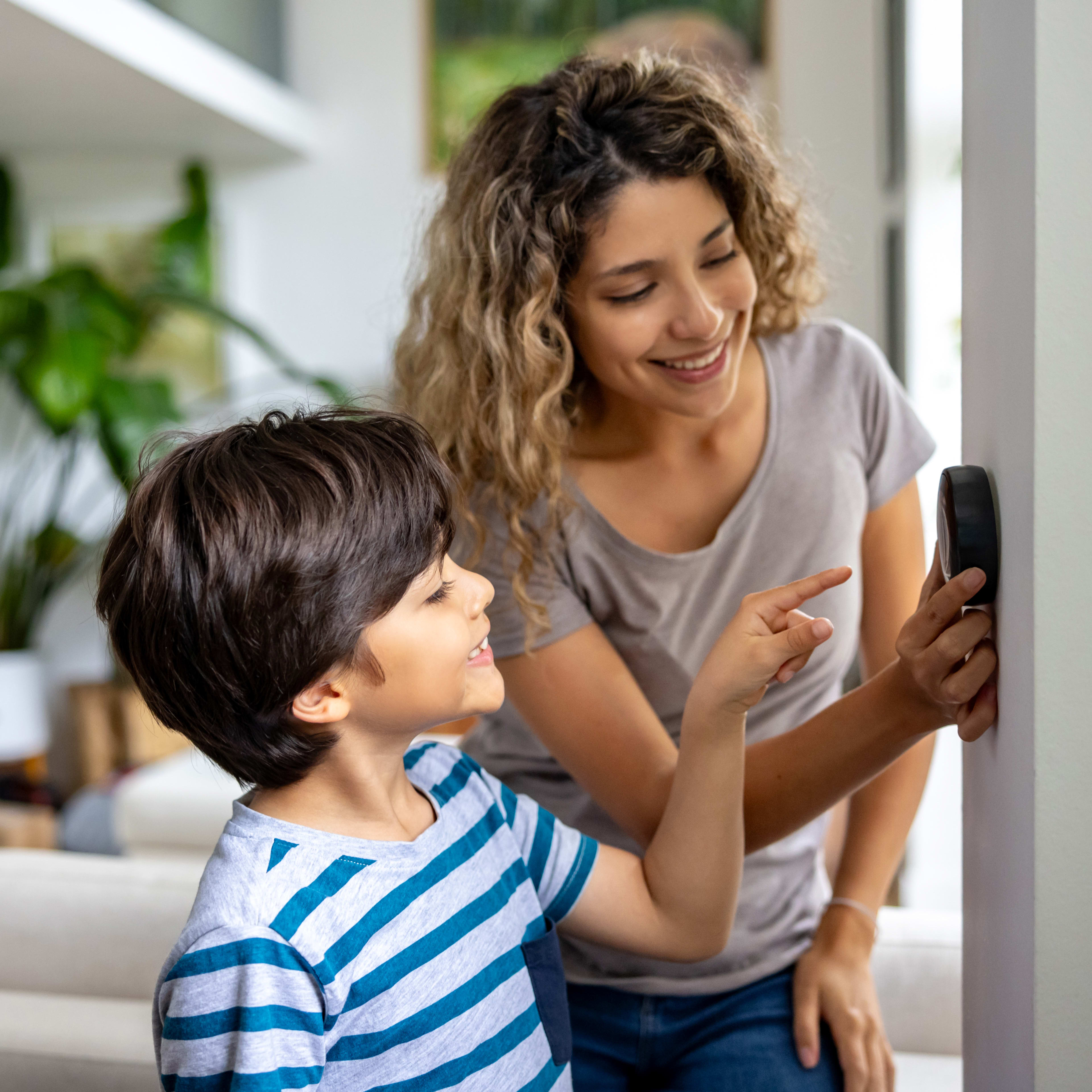 A young woman with curly hair is smiling and interacting with a young boy wearing a striped shirt in what appears to be a cozy, indoor living space with potted plants visible in the background.