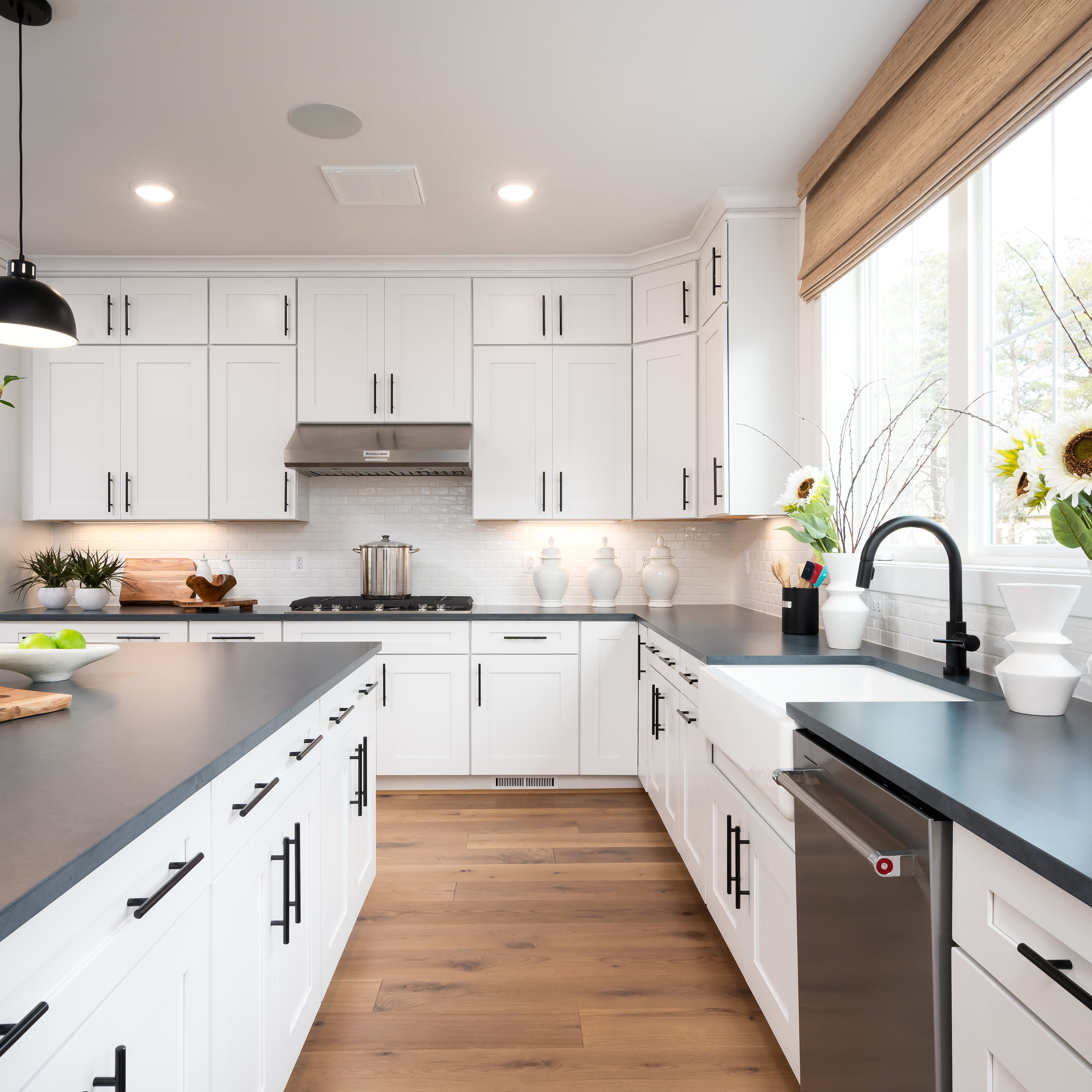 A modern, bright kitchen with white cabinets, gray countertops, and wooden floors, featuring a large window that lets in natural light and showcases a vase of sunflowers on the windowsill.