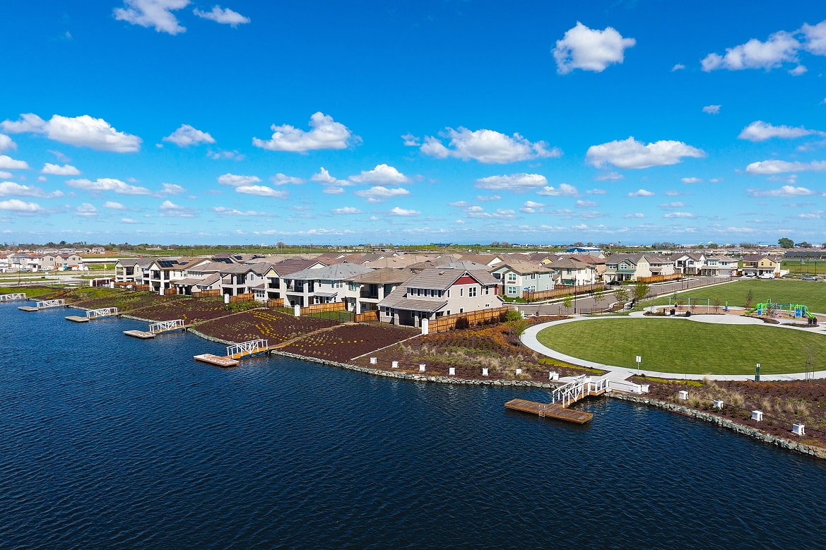 A scenic waterfront community with colorful houses, a grassy park, and a clear blue sky with fluffy white clouds.