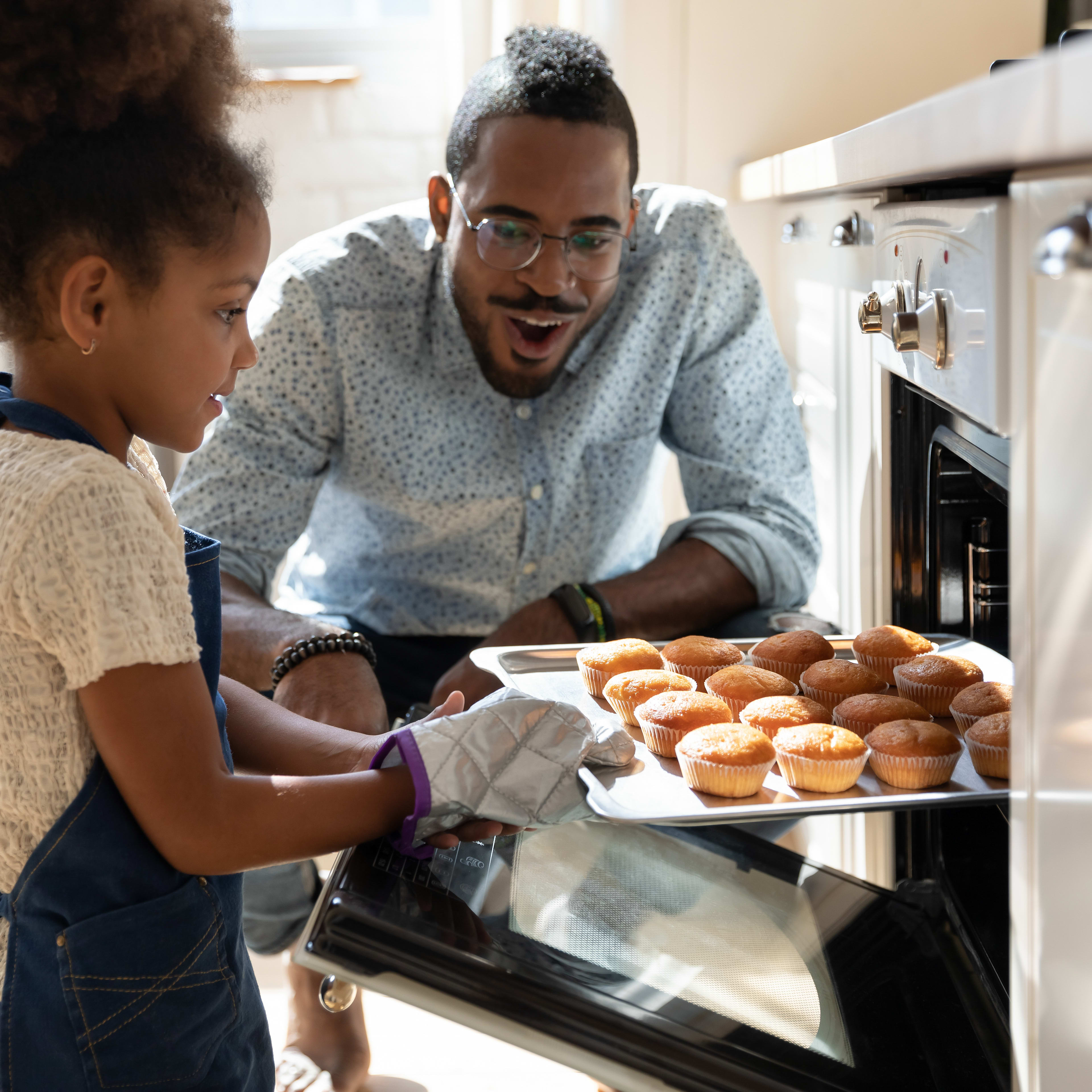 A man in a gray sweater is smiling and holding a tray of cupcakes as he interacts with a young girl in the kitchen.