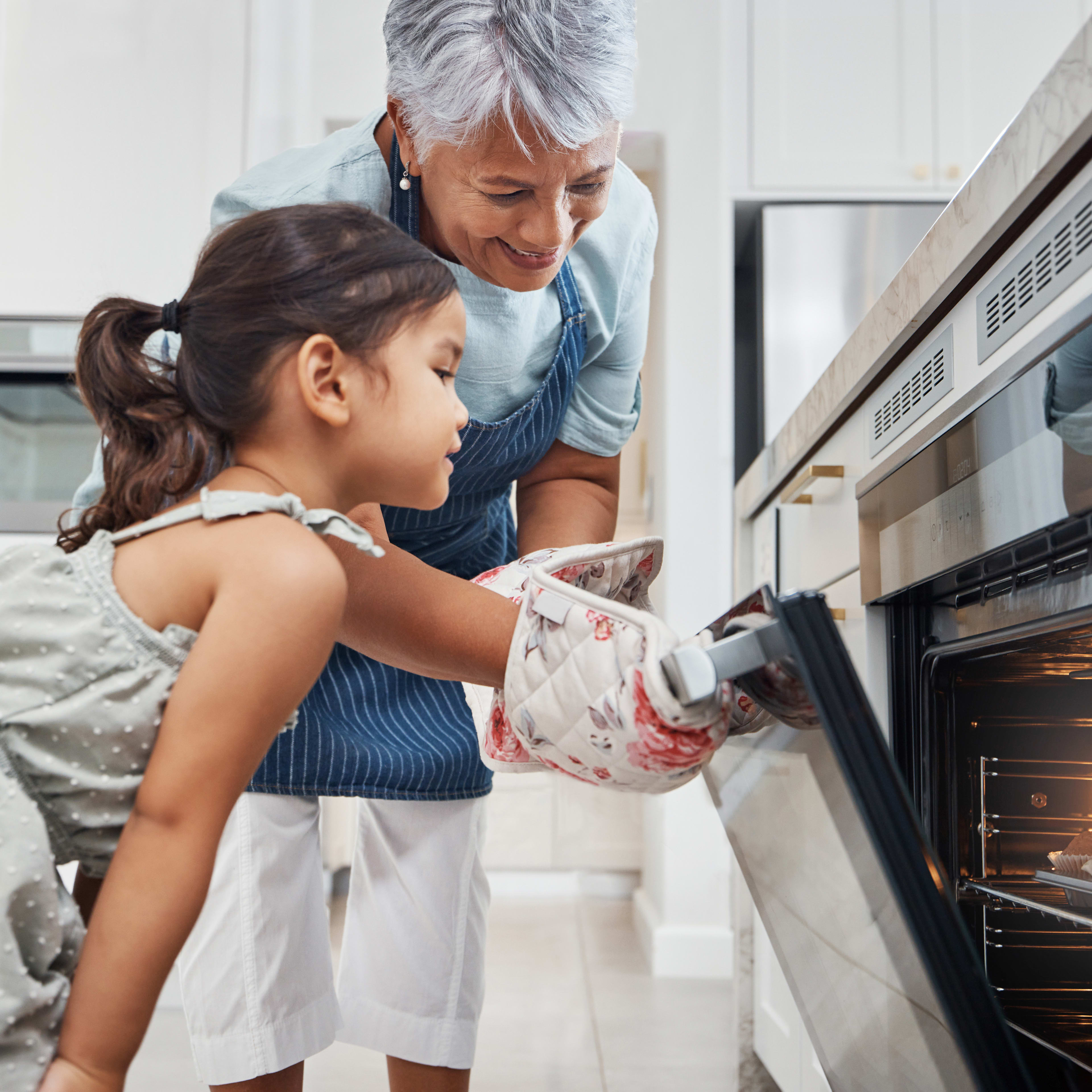 A young girl in a floral dress is standing in a kitchen, with an older adult, likely a parent or guardian, assisting her as she opens the oven door.