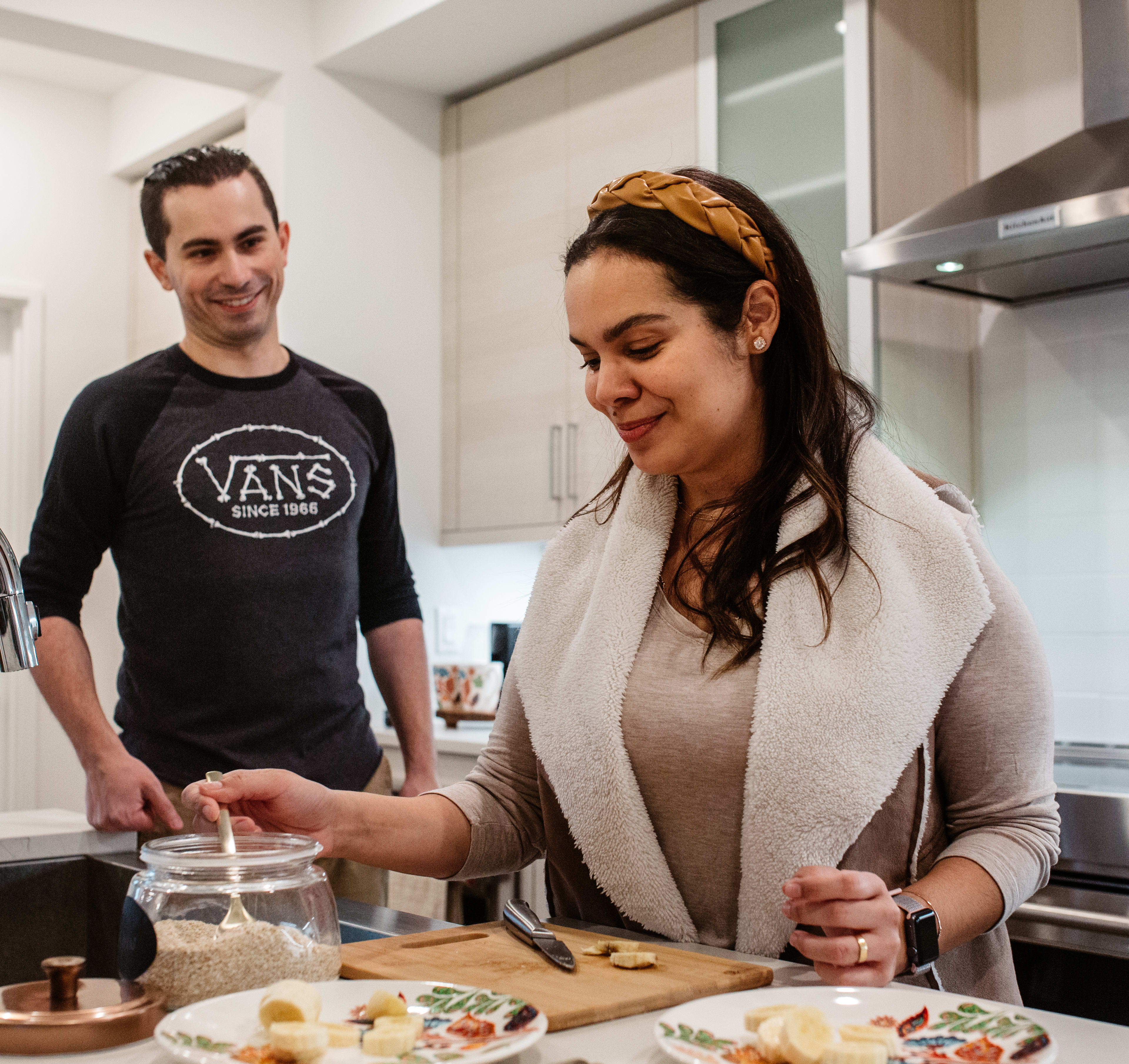 A young couple is preparing food together in a modern kitchen, with the woman wearing a headband and the man wearing a Vans t-shirt.