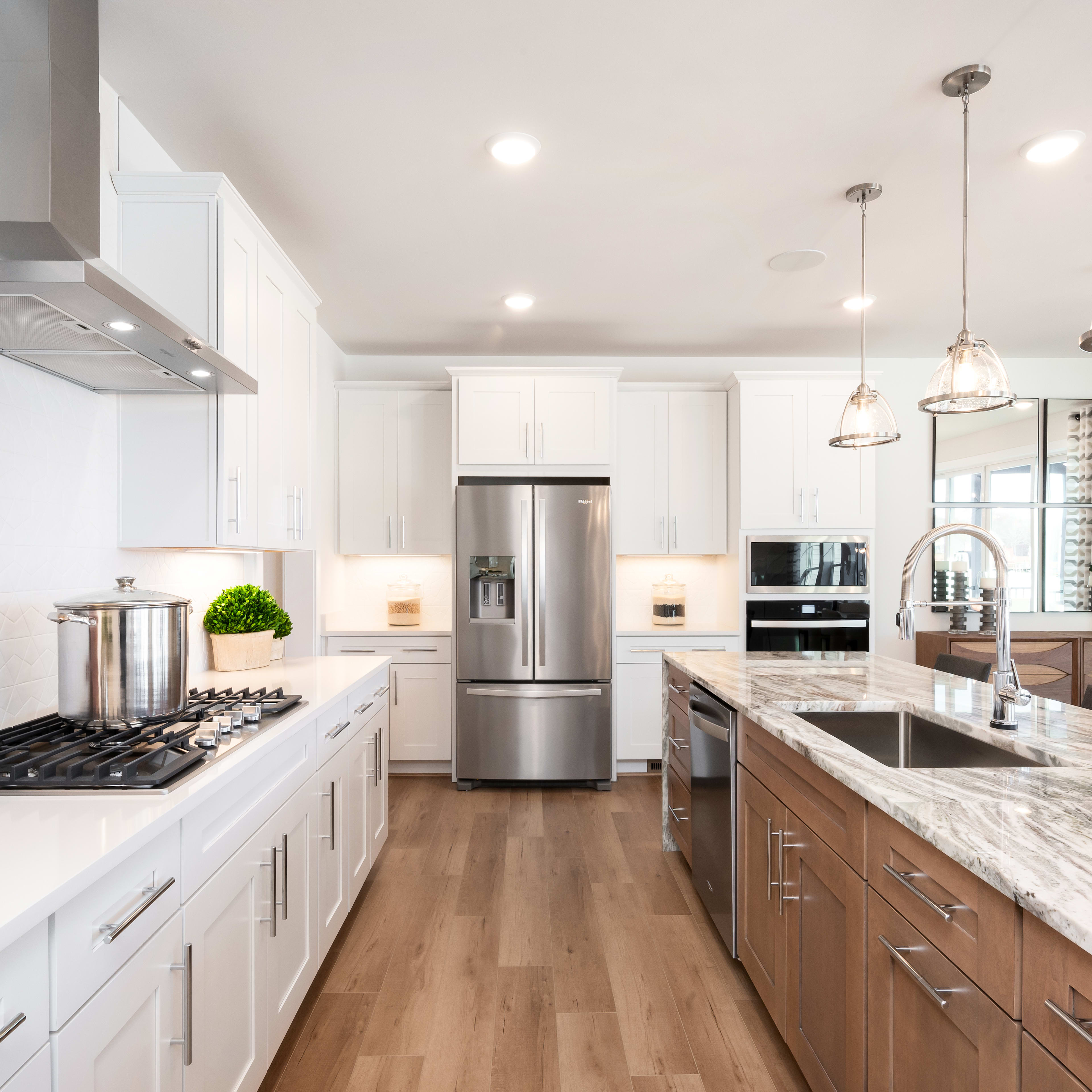 A modern, well-lit kitchen with white cabinets, marble countertops, and stainless steel appliances, featuring a long central island and pendant lighting.