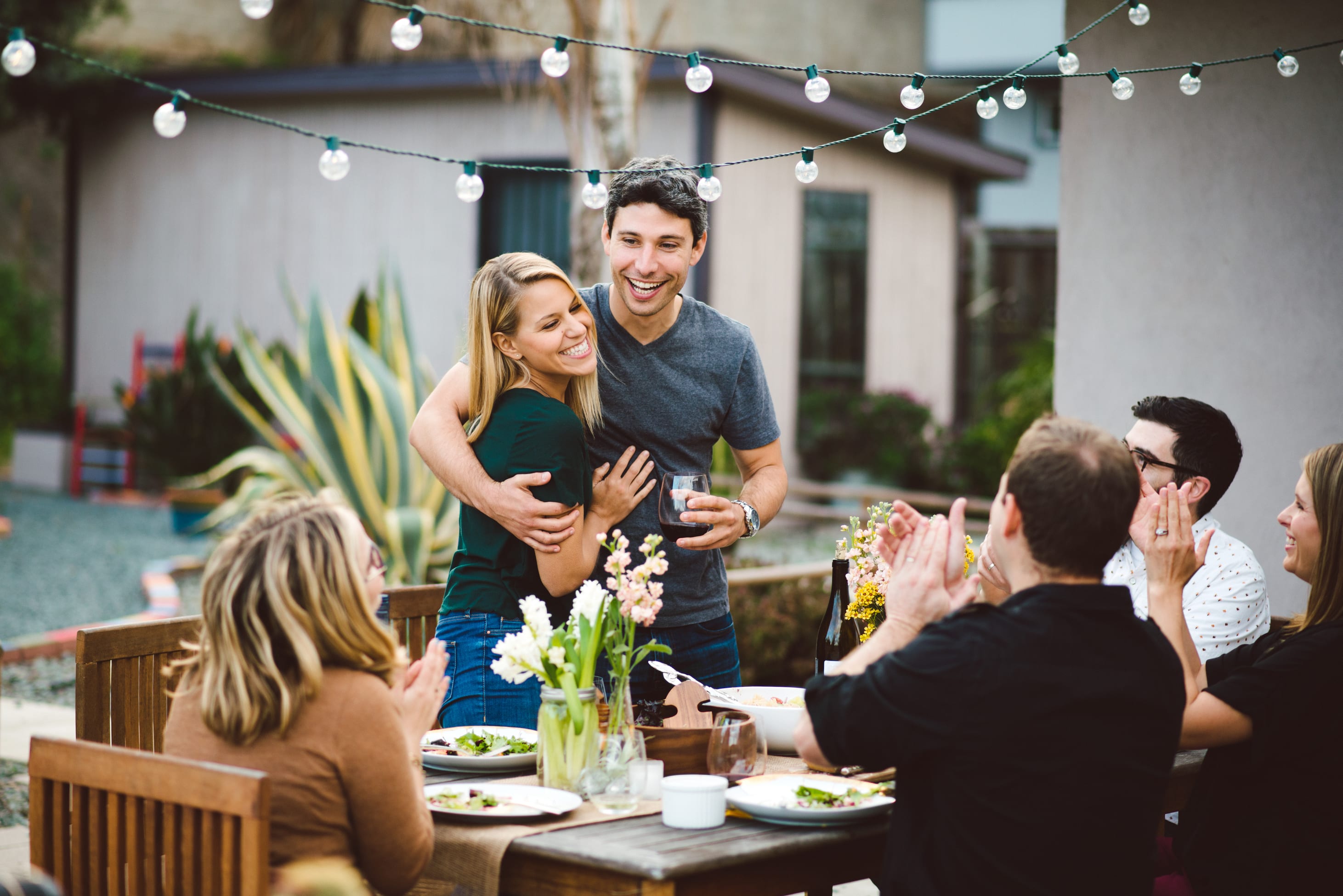 A group of people enjoying an outdoor gathering, with a couple embracing in the foreground and string lights overhead, surrounded by a lush, natural setting.
