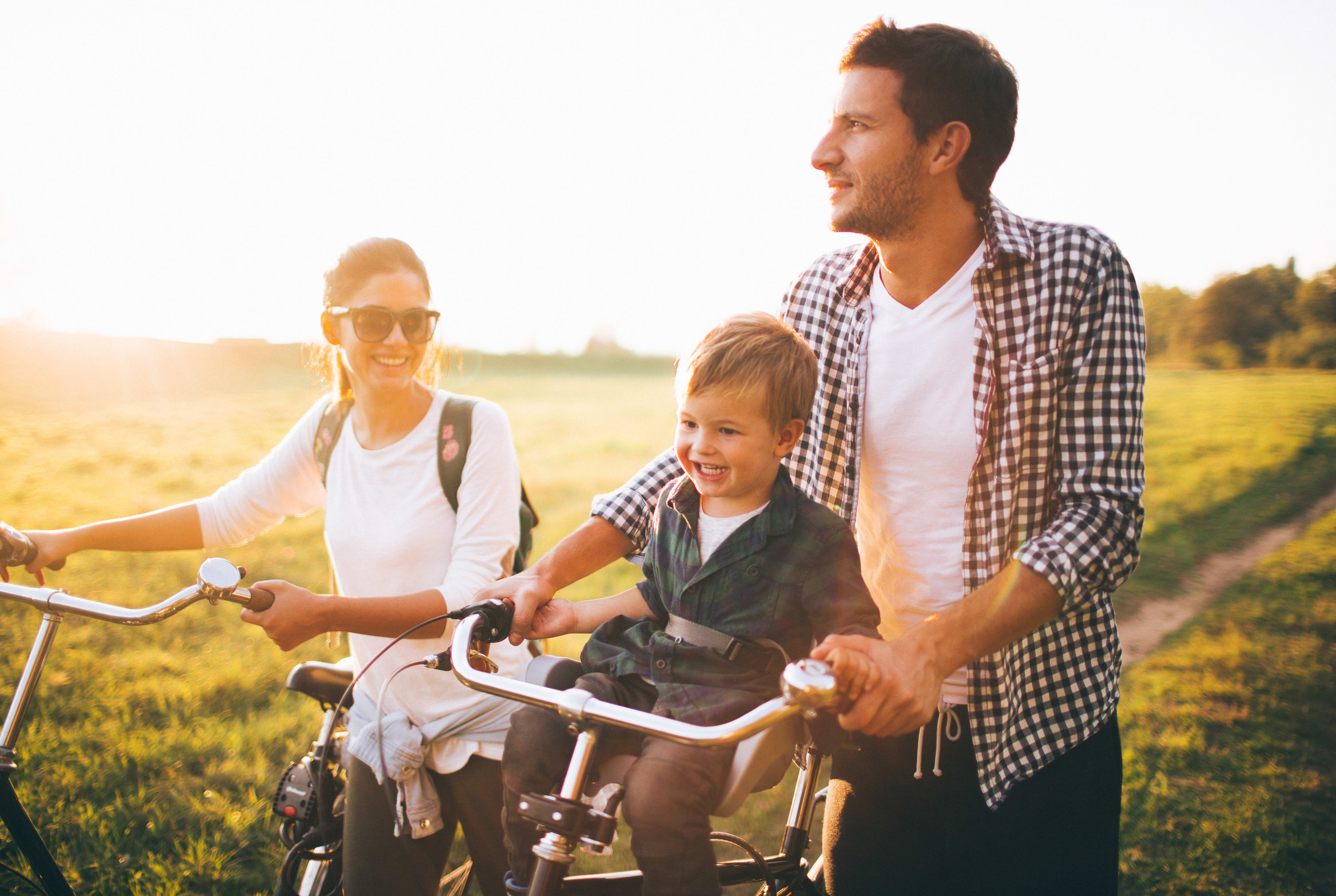 A family of three, a man, a woman, and a young child, riding bicycles together through a scenic, sun-drenched field.