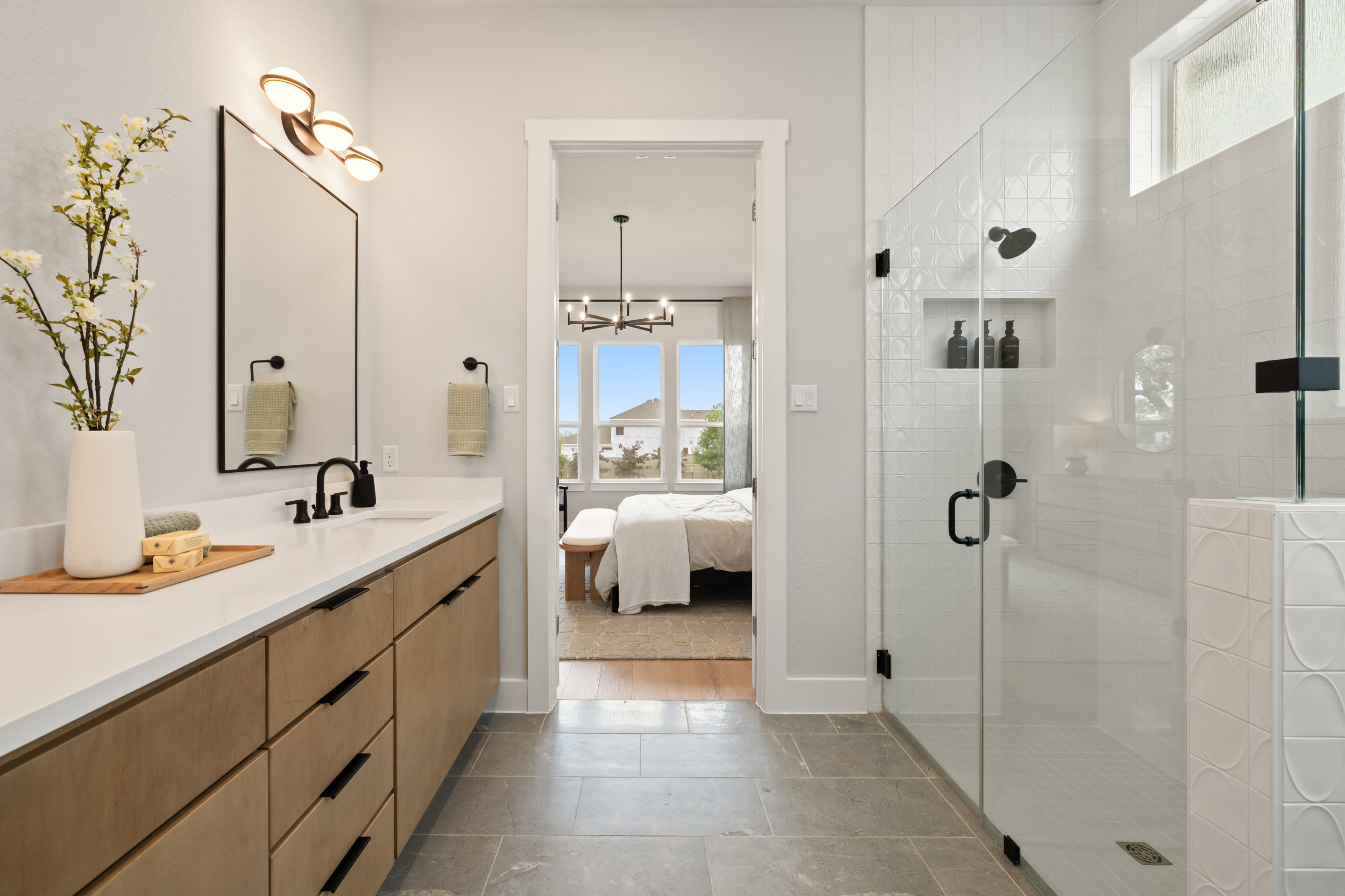 A modern and minimalist bathroom with a wooden vanity, a large mirror, and a glass-enclosed shower, leading to a bedroom with a window and natural light.