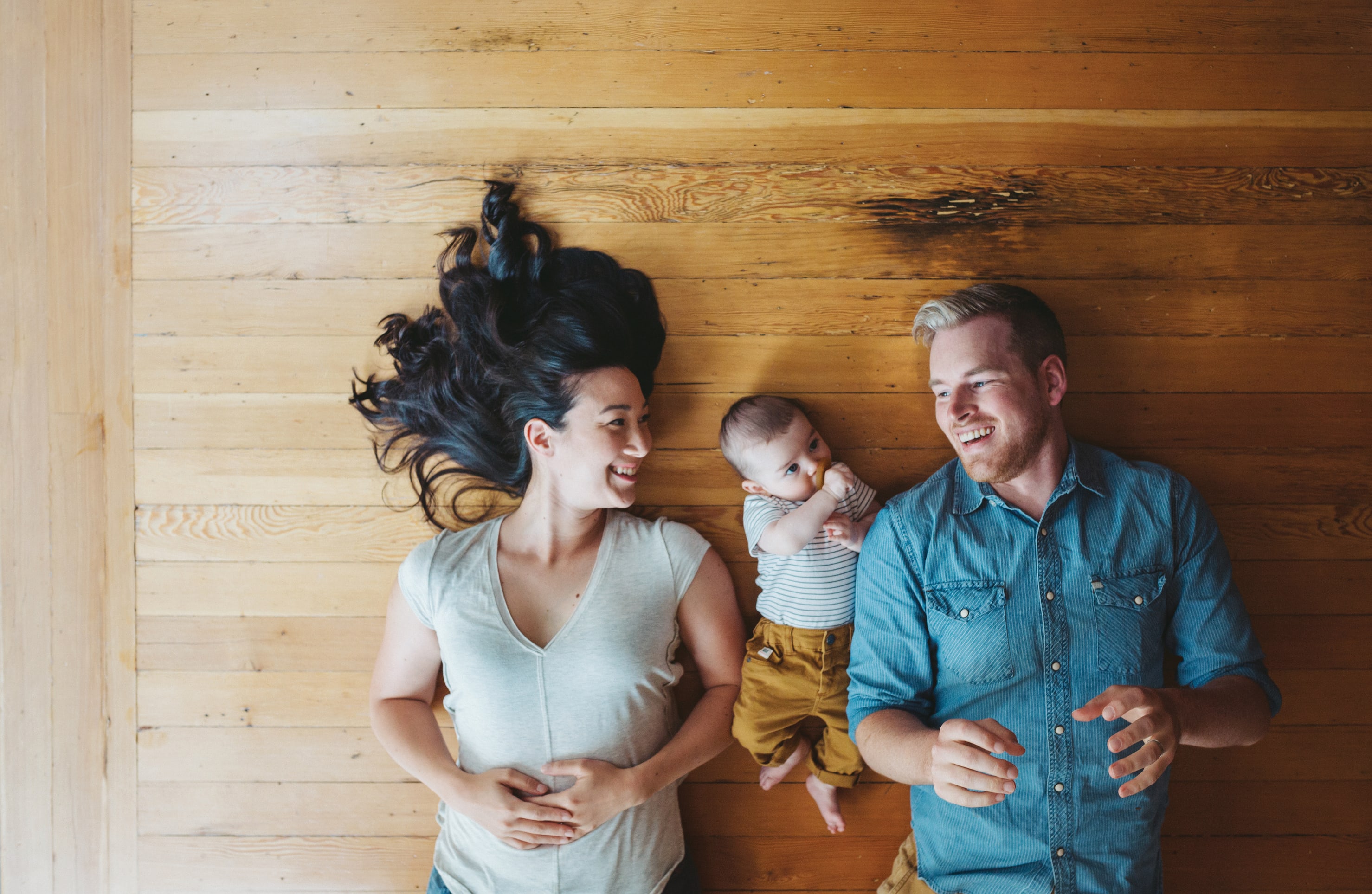A young family, consisting of a smiling woman, a man, and a baby, stands in front of a wooden wall backdrop.