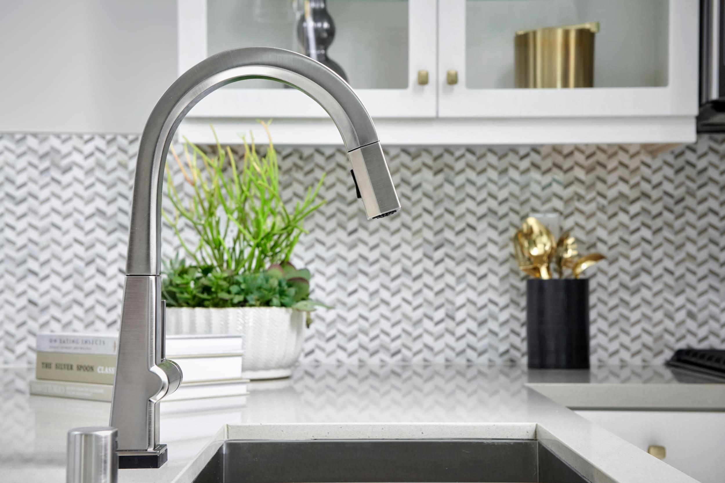 A modern kitchen sink with a sleek faucet, surrounded by a herringbone tile backsplash and accented by potted plants and decorative elements.