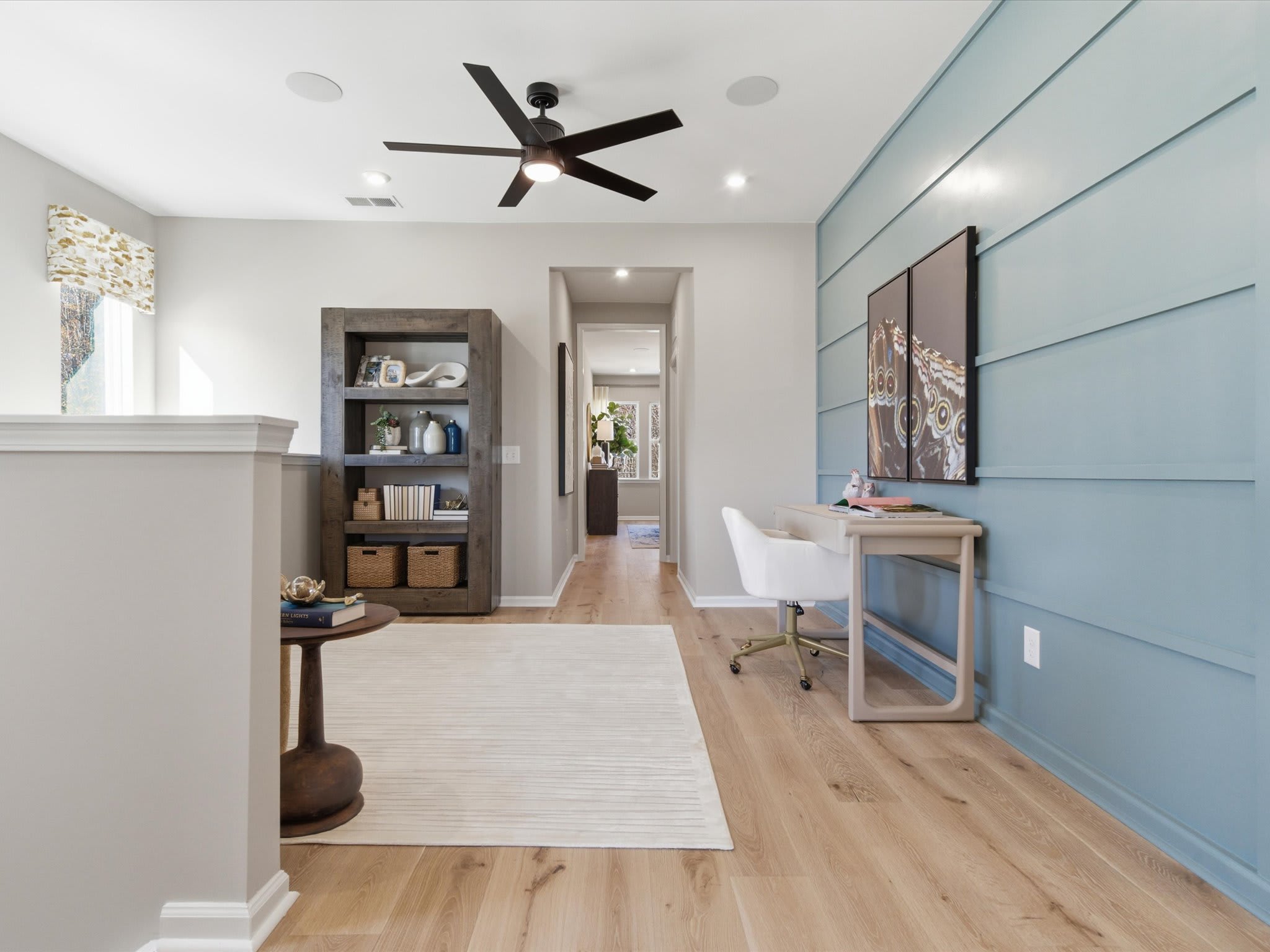 A spacious and well-lit home office with a ceiling fan, wooden floors, and a mix of modern and rustic decor elements, including a bookshelf, a desk, and a wall-mounted artwork.