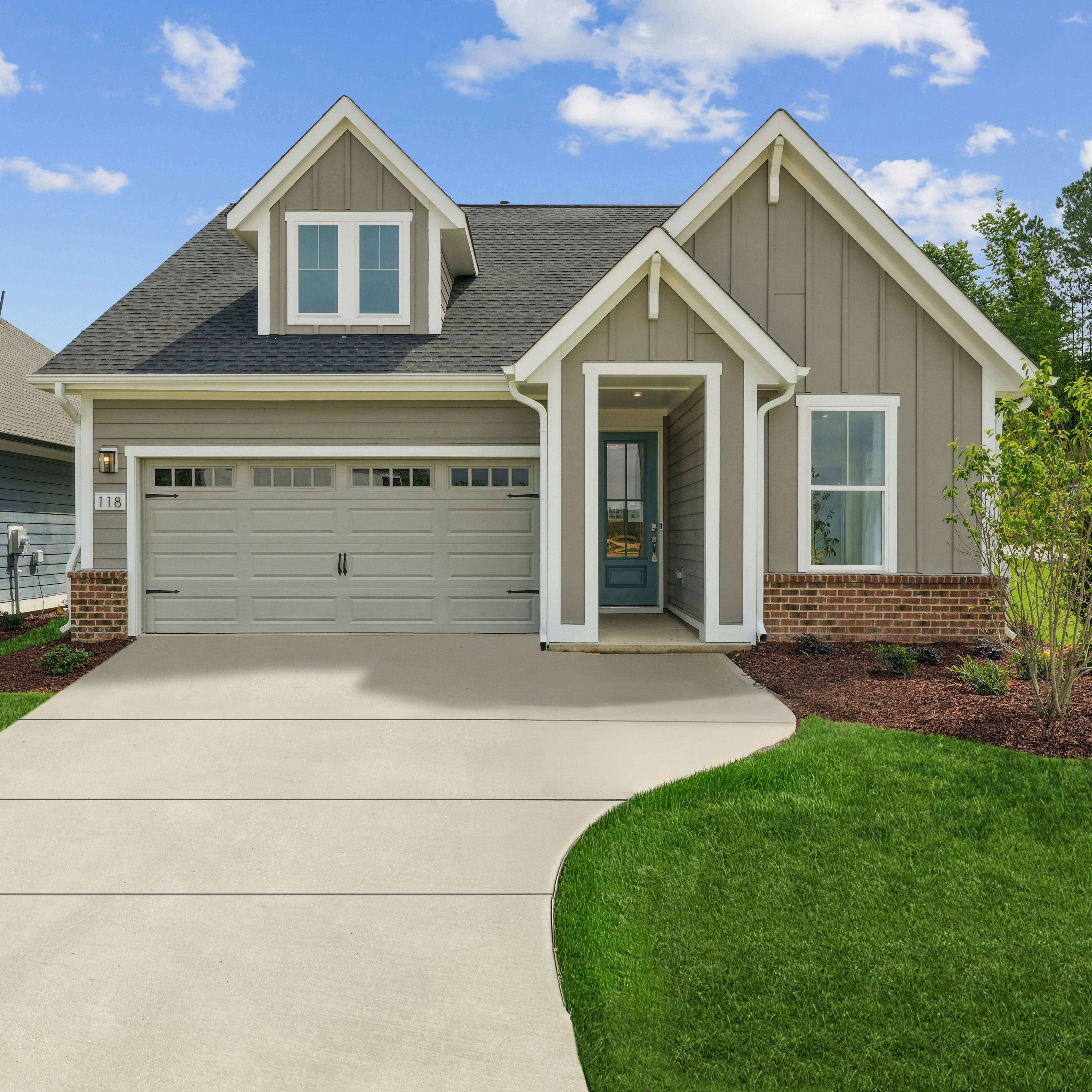 A well-manicured lawn leads to a two-story house with a gray exterior, a garage, and a lush green landscape in the background.