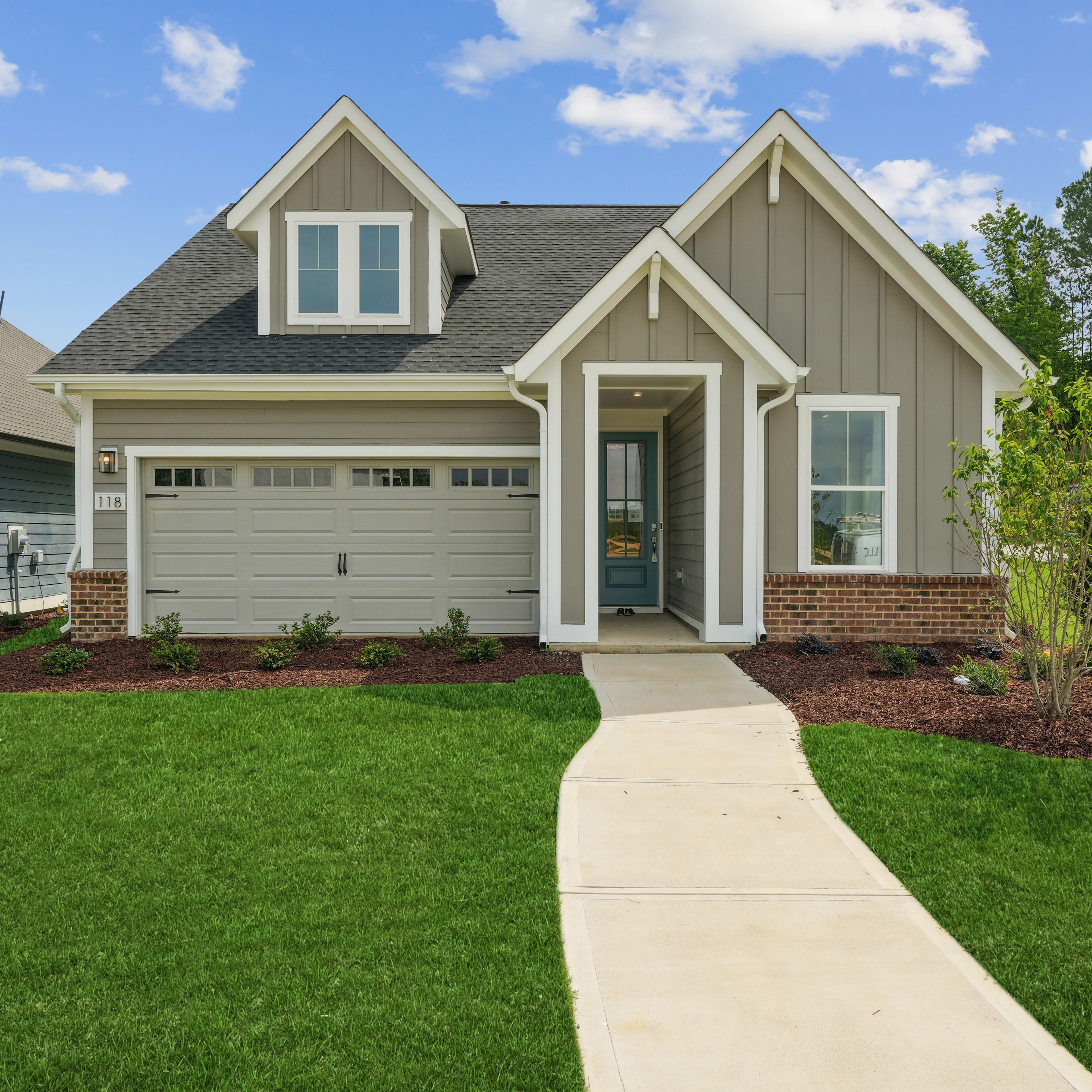 A well-manicured lawn leads to a two-story house with a gabled roof, siding in a neutral color, and a garage door, set against a backdrop of blue sky with fluffy white clouds and lush green trees.