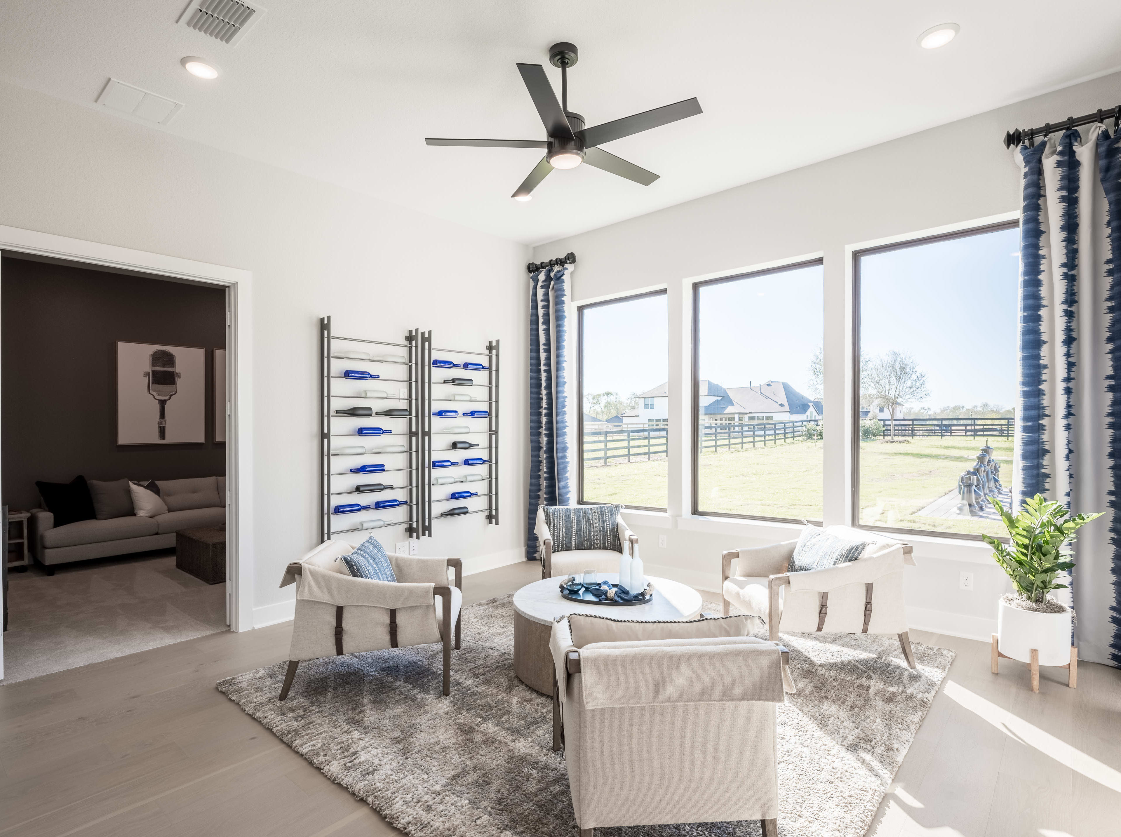 A spacious and well-lit living room with a ceiling fan, large windows overlooking a scenic outdoor area, and comfortable seating arrangements including armchairs and a sofa.
