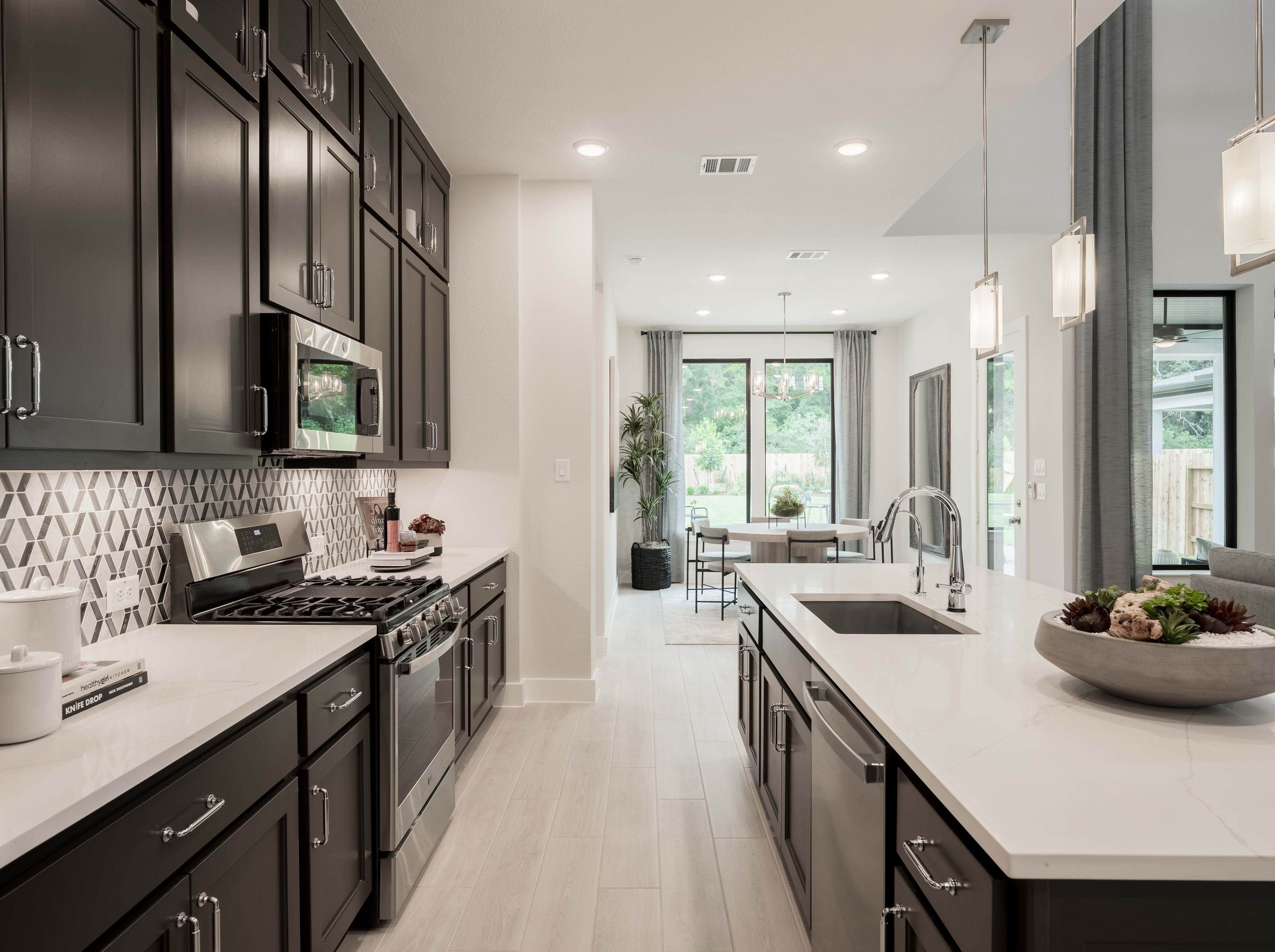 A modern, sleek kitchen with dark cabinets, white countertops, and a large window overlooking a natural setting.