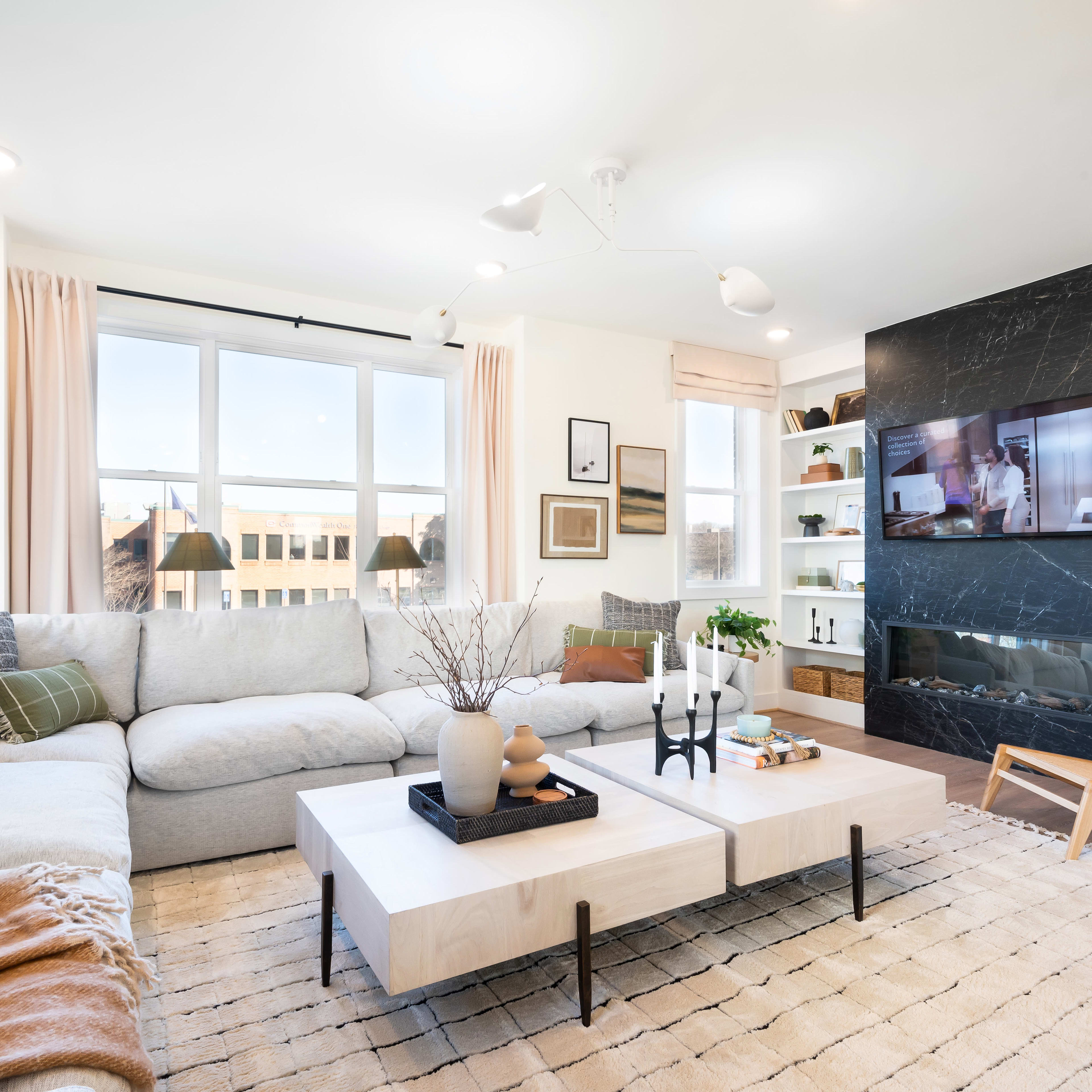 A cozy and inviting living room with a large sectional sofa, a coffee table, and a wall-mounted TV surrounded by shelves and decor, all set against a backdrop of large windows overlooking an outdoor area.