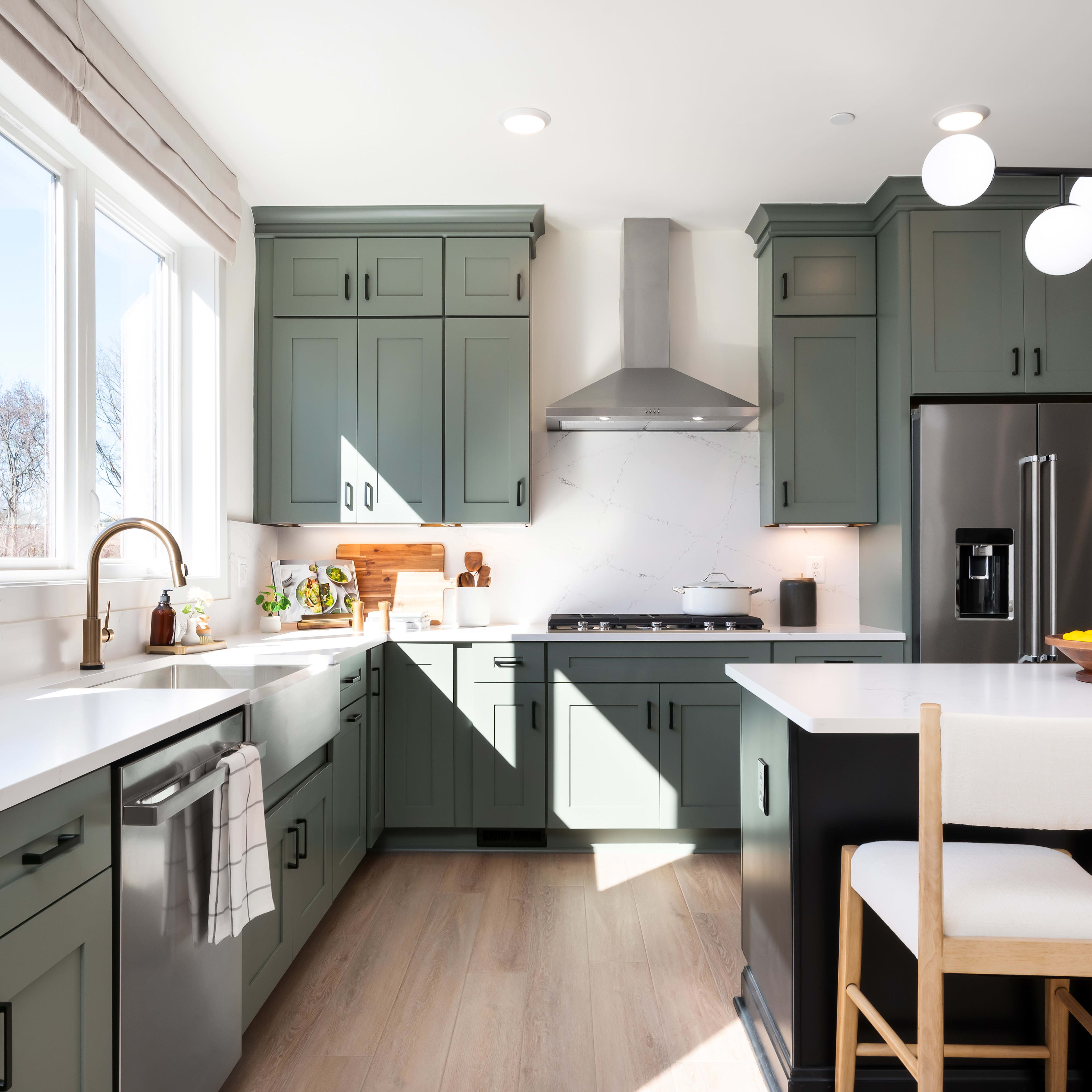 A modern, well-lit kitchen with green cabinets, white countertops, and a dining area with stools in the foreground.