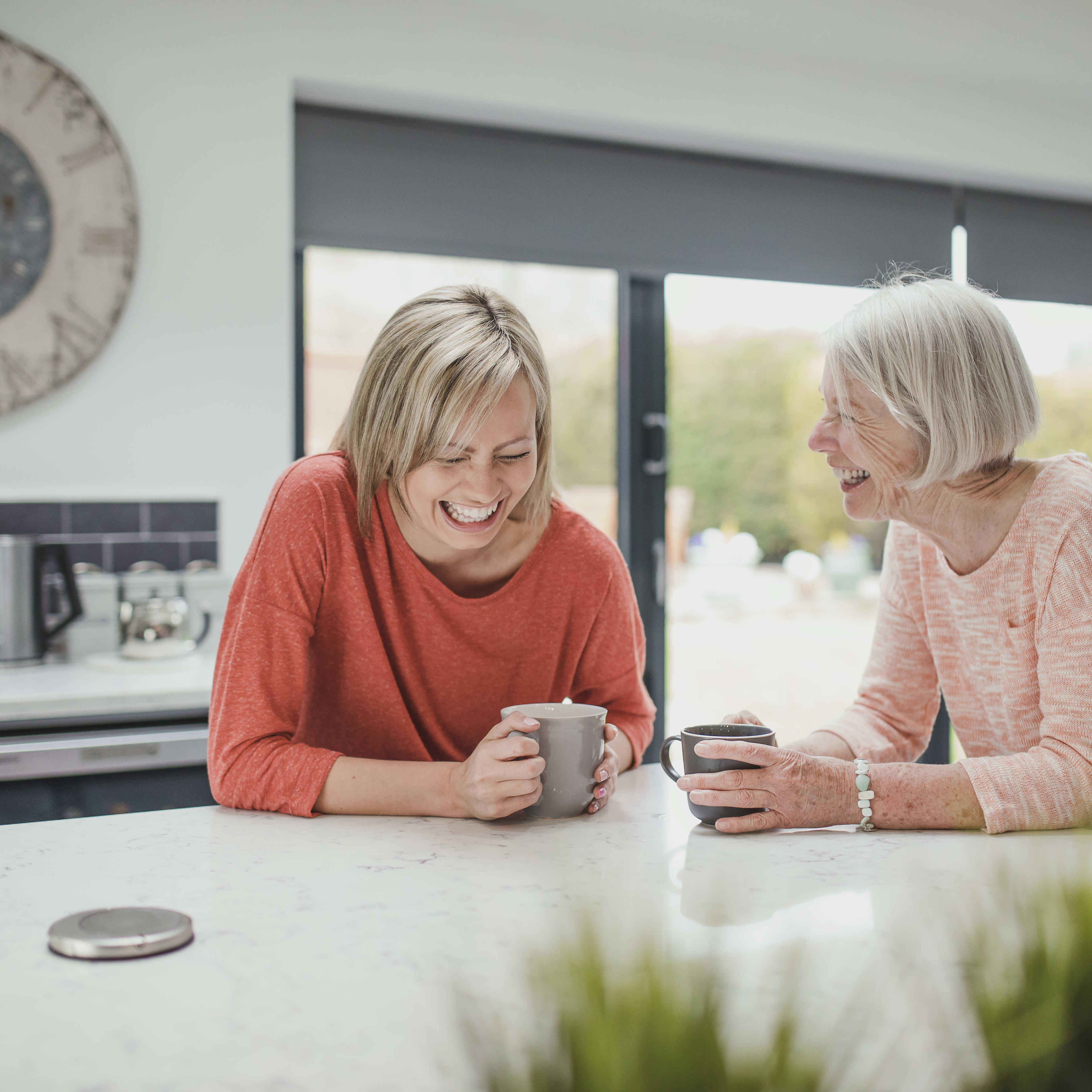 Two women, one wearing a red sweater and the other a pink sweater, are sitting at a kitchen counter and enjoying a conversation while holding mugs.