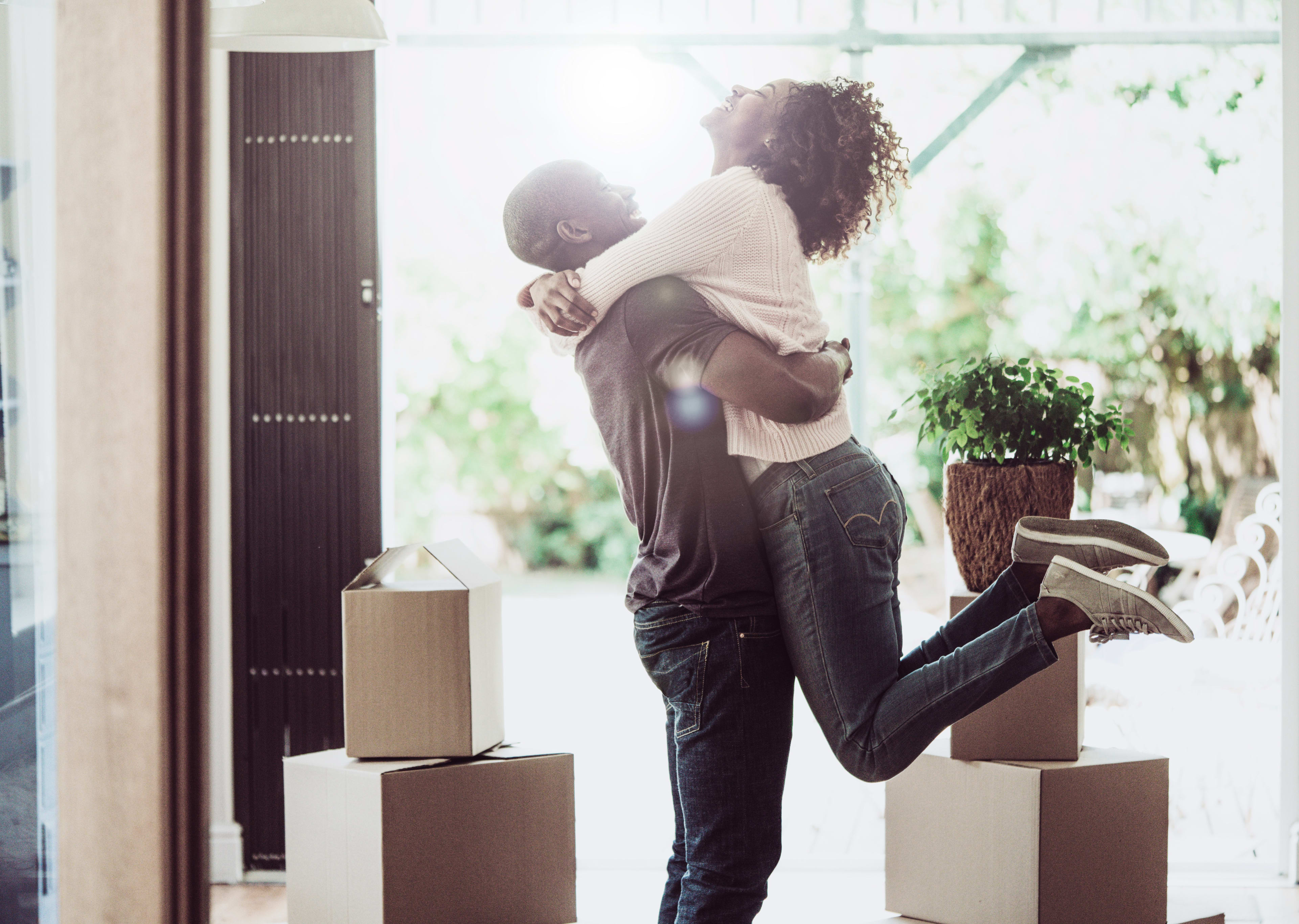 A couple embracing in a room surrounded by moving boxes, with a window in the background providing natural lighting.