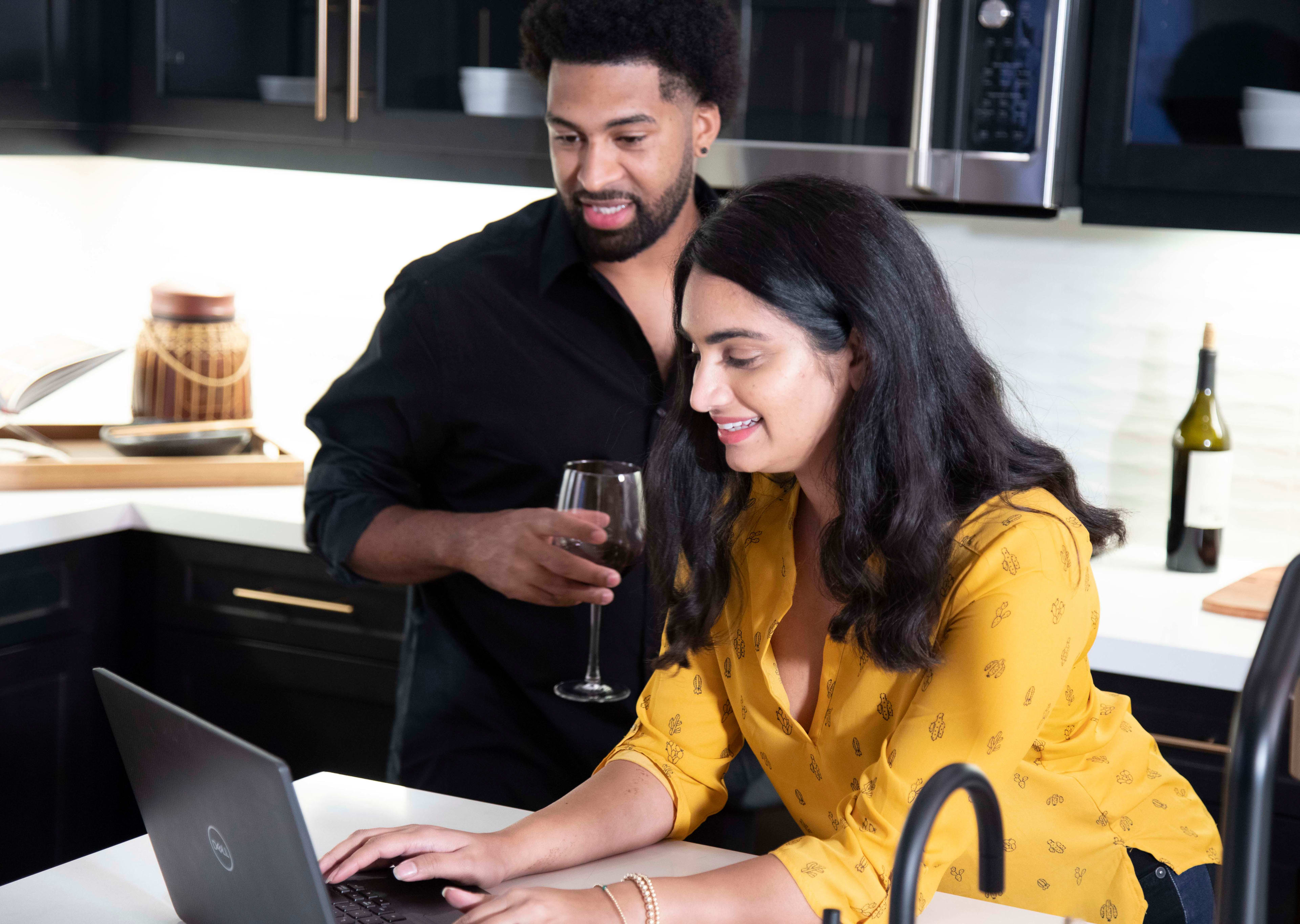 A man in a black shirt is embracing a woman in a yellow top as they sit together in a kitchen, with various kitchen appliances and utensils visible in the background.